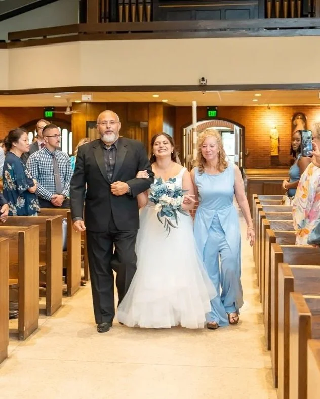 Bride walking down the aisle with her father and a woman, surrounded by guests in a church setting.