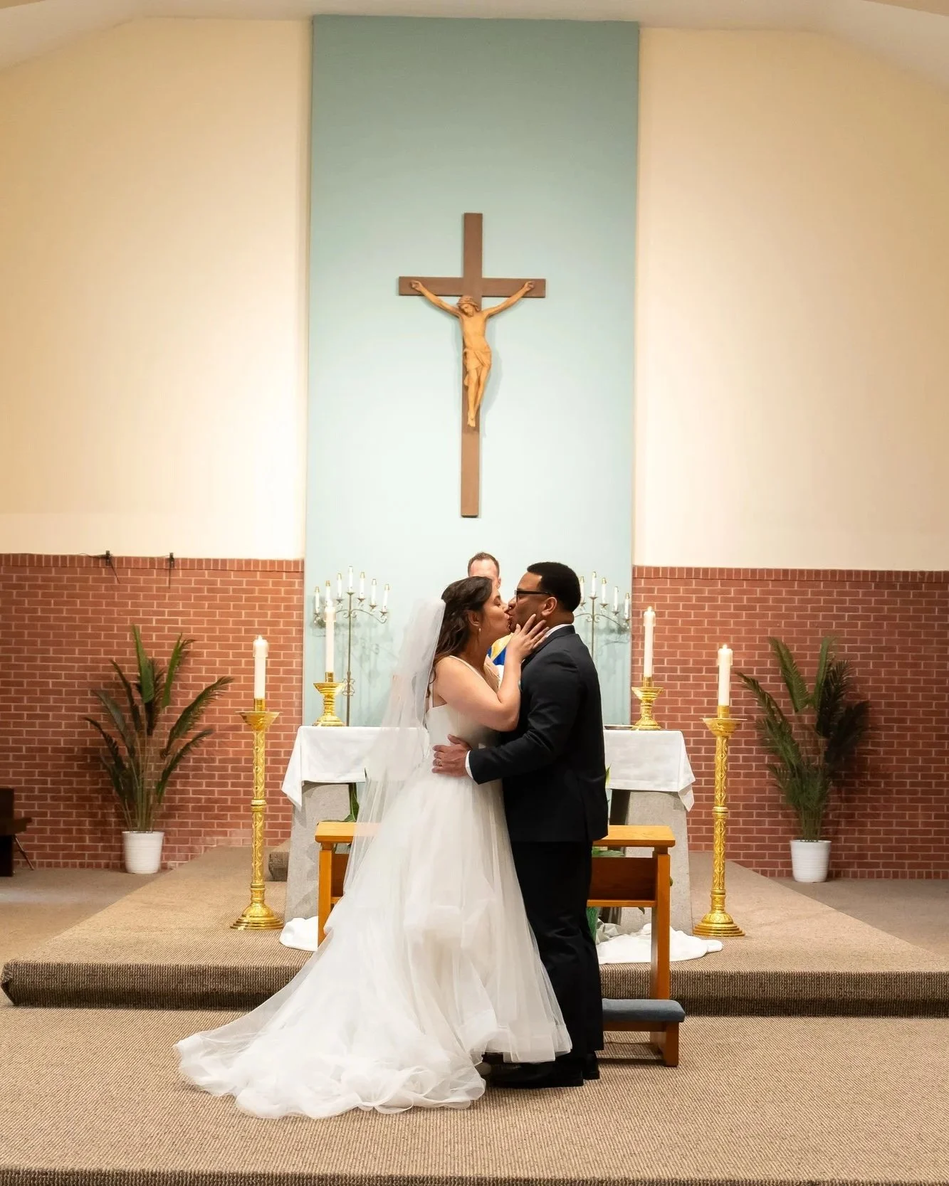 A couple kissing during a wedding ceremony in a church with a large crucifix on the wall behind them, lit candles, and potted plants.