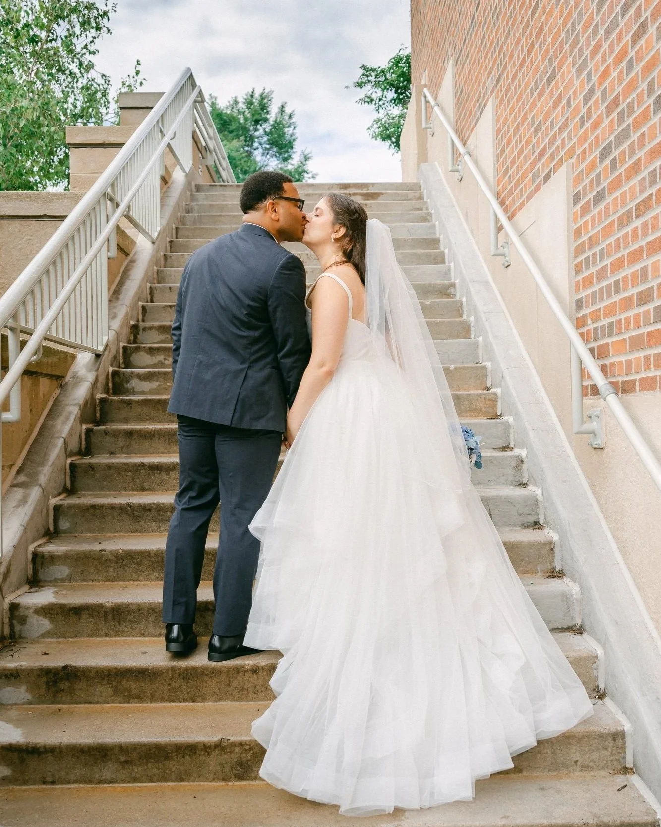 A newlywed couple kissing on outdoor stairs, with the bride in a white wedding dress and veil, and the groom in a dark suit, during daytime.