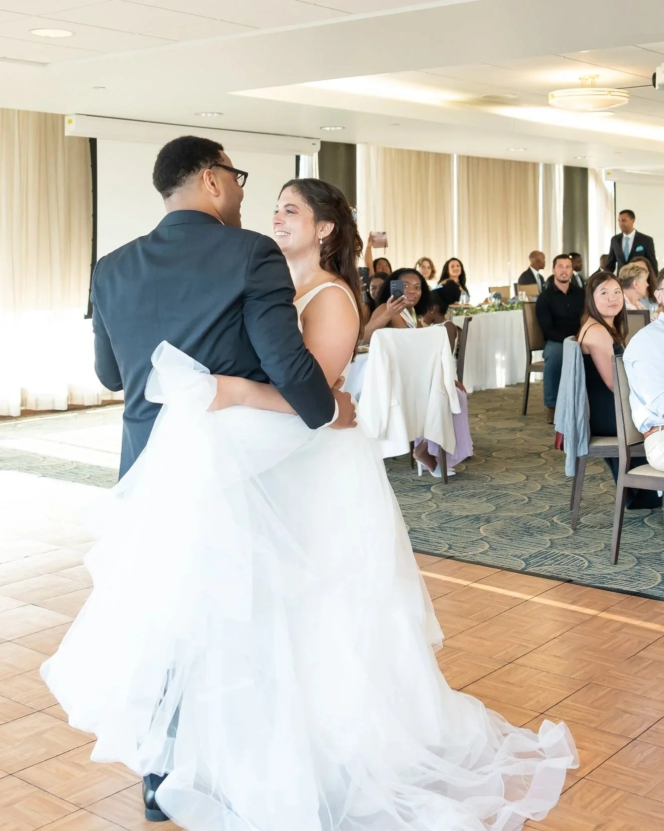 A newlywed couple dancing at their wedding reception, with guests seated at tables watching and taking photos.