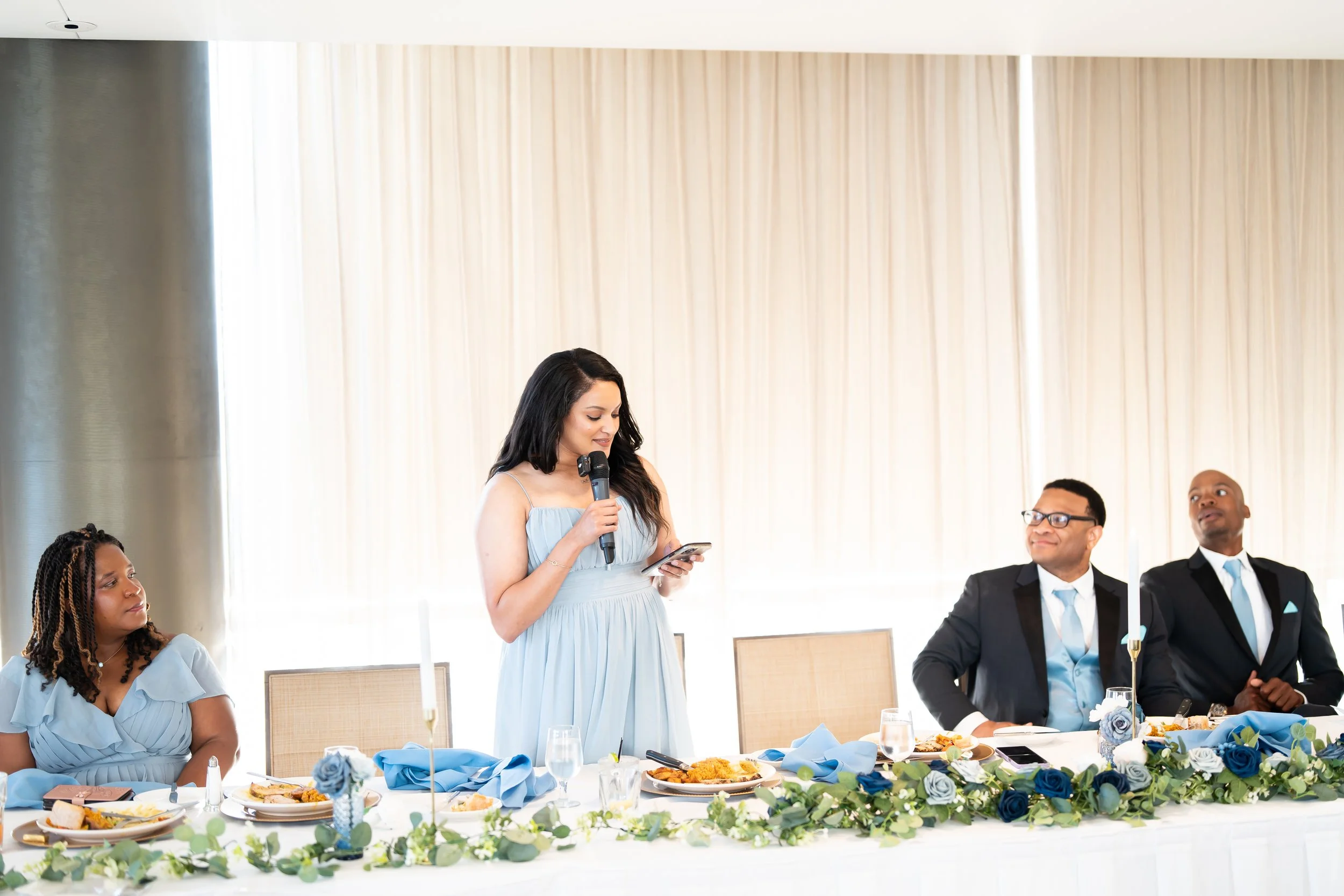 A woman in a light blue dress giving a speech at a table with four seated people during a formal event.