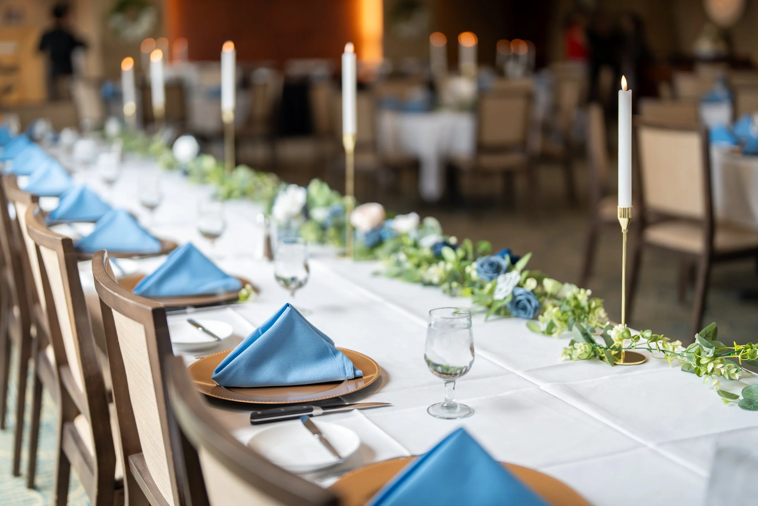 Elegant banquet table setup with blue napkins, white tablecloth, floral centerpiece, gold candlesticks, and glassware.