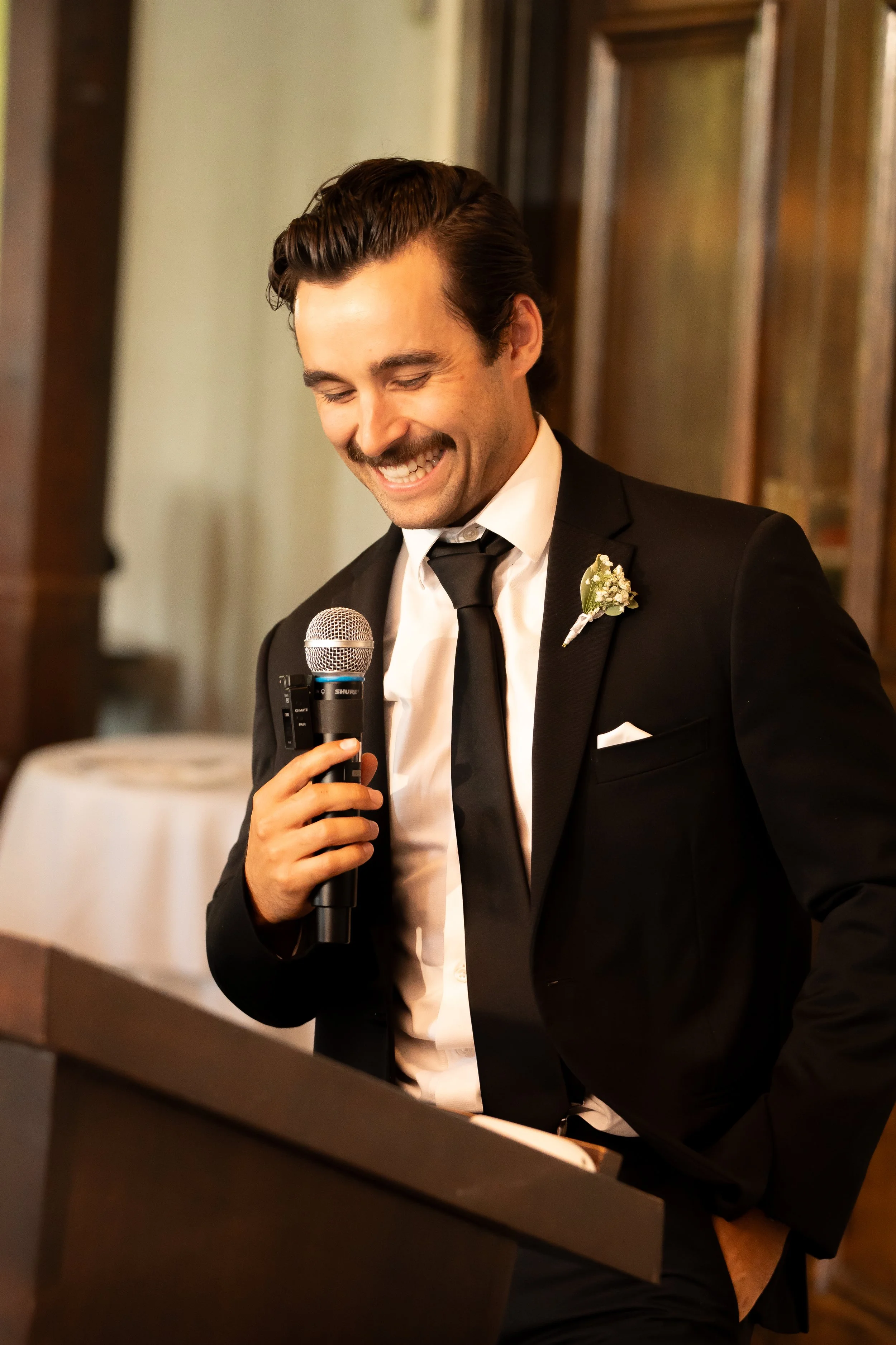 A man in a black suit and tie smiling while holding a microphone, giving a speech at a formal event.
