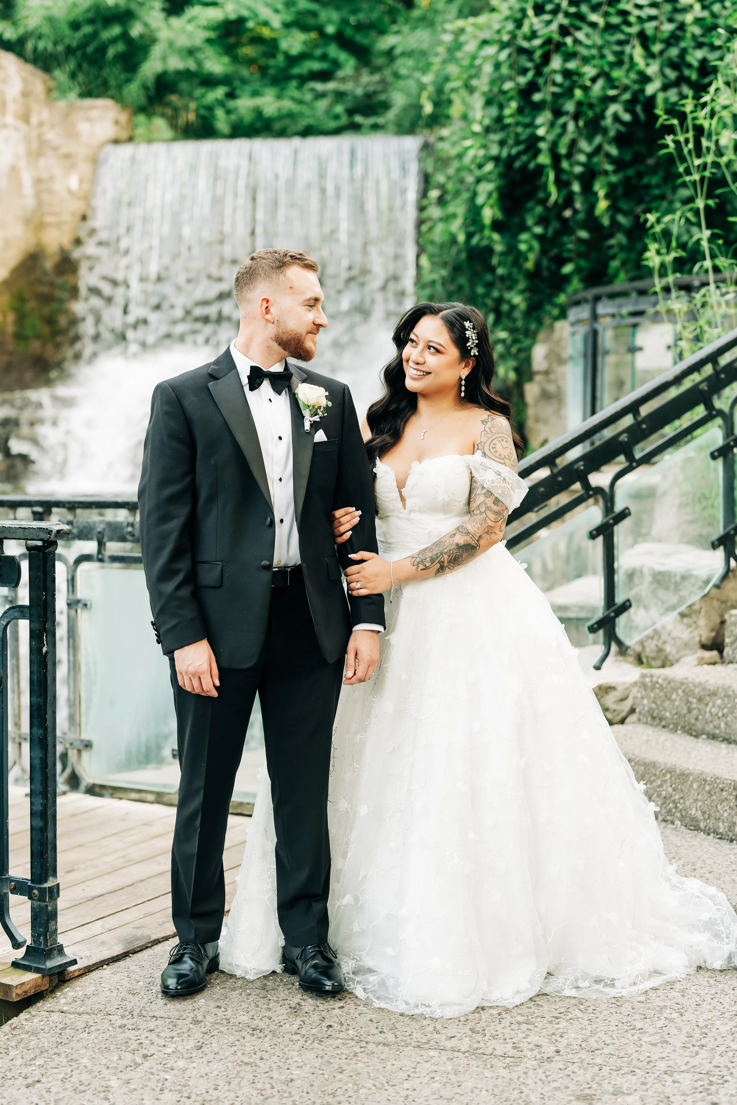 A bride and groom standing outdoors near a waterfall, gazing at each other. The groom is wearing a black tuxedo with a bow tie, and the bride is in a white wedding gown with lace details and tattoos on her arms.