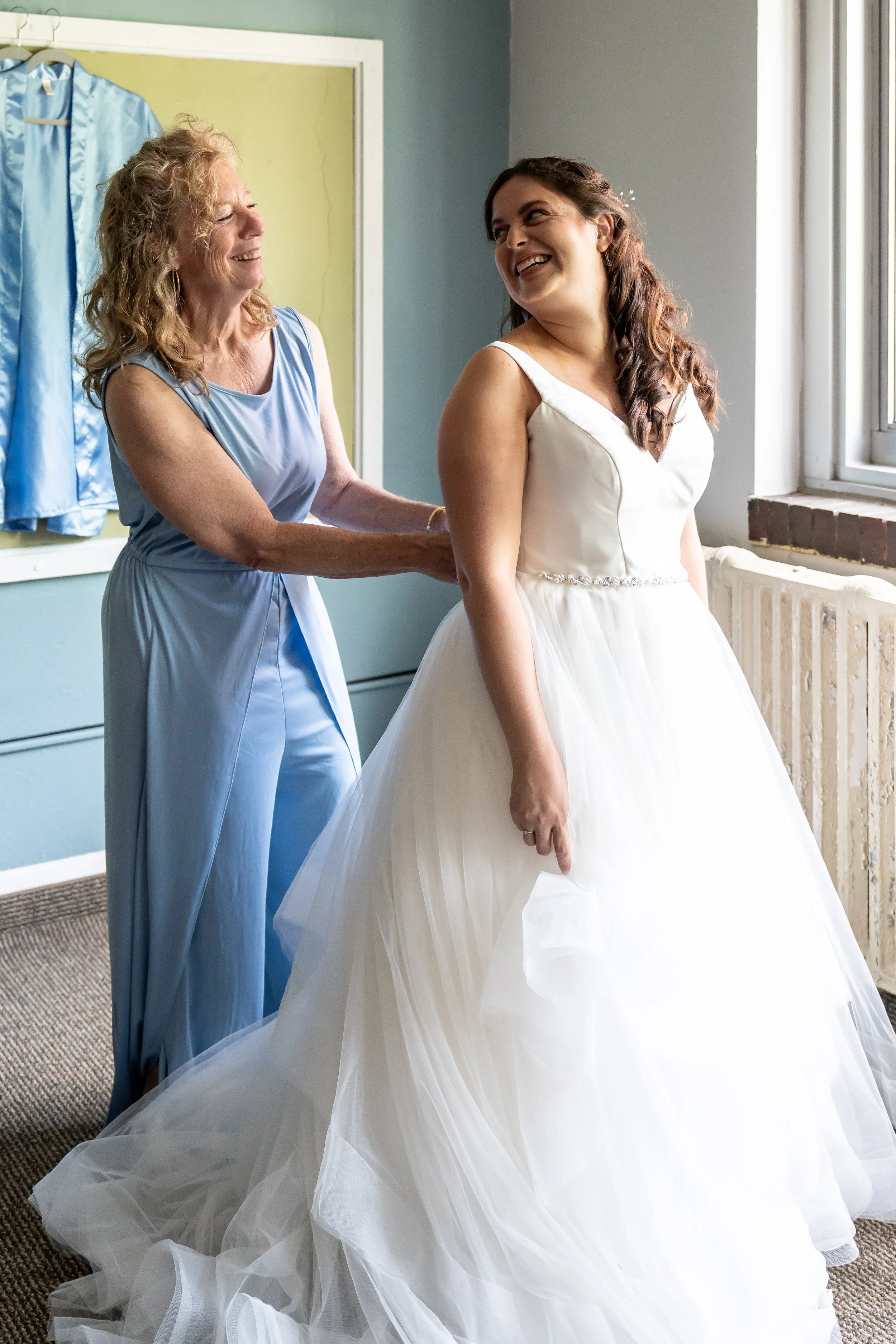 A bride in a white wedding dress smiling and adjusting her gown, while an older woman in a blue dress helps her, in a room with a window and a blue garment hanging on the wall.