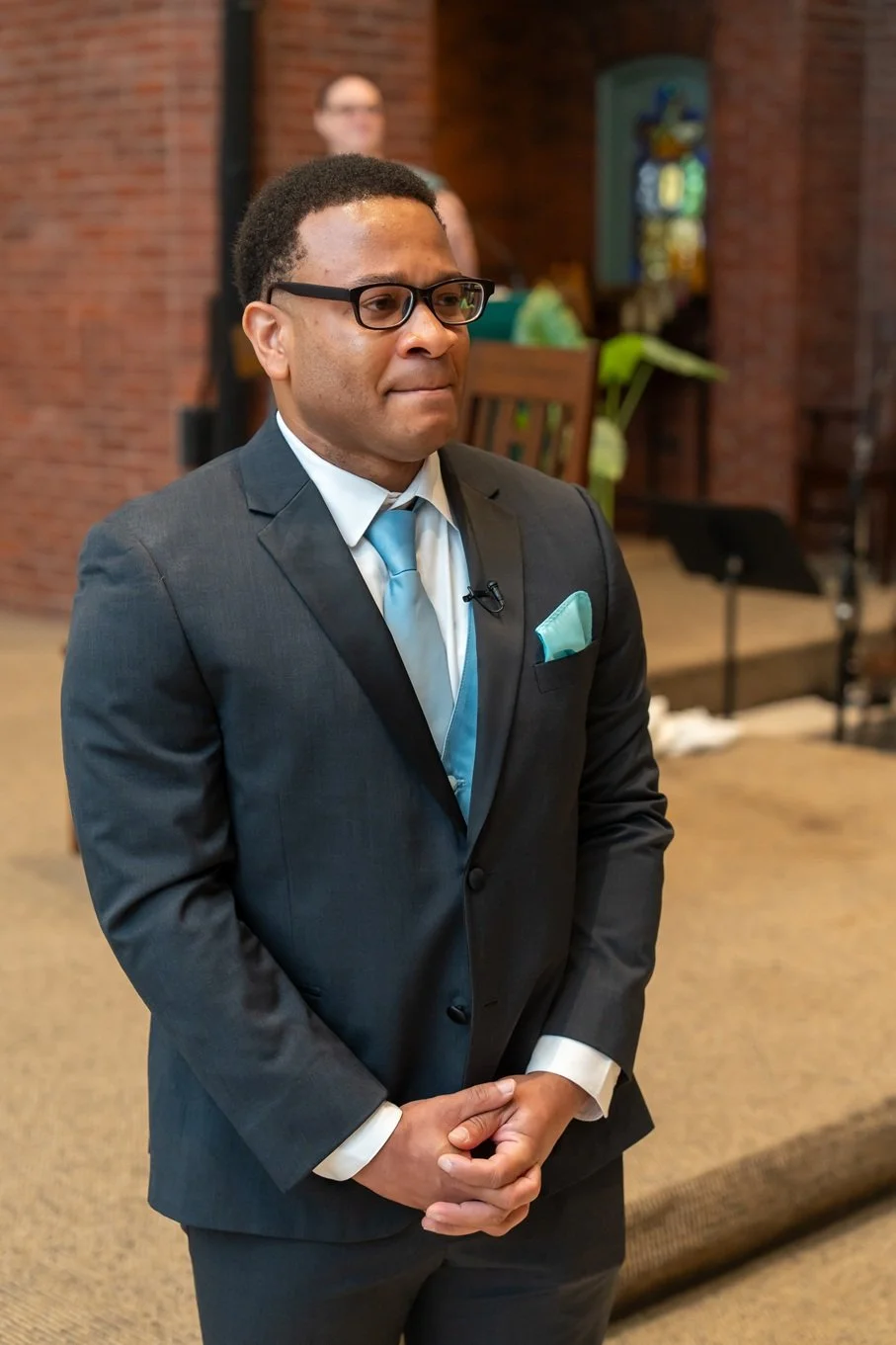 A man in a dark suit with a light blue tie and pocket square standing in a church. He is wearing glasses and appears emotional or thoughtful, with his hands clasped in front of him.