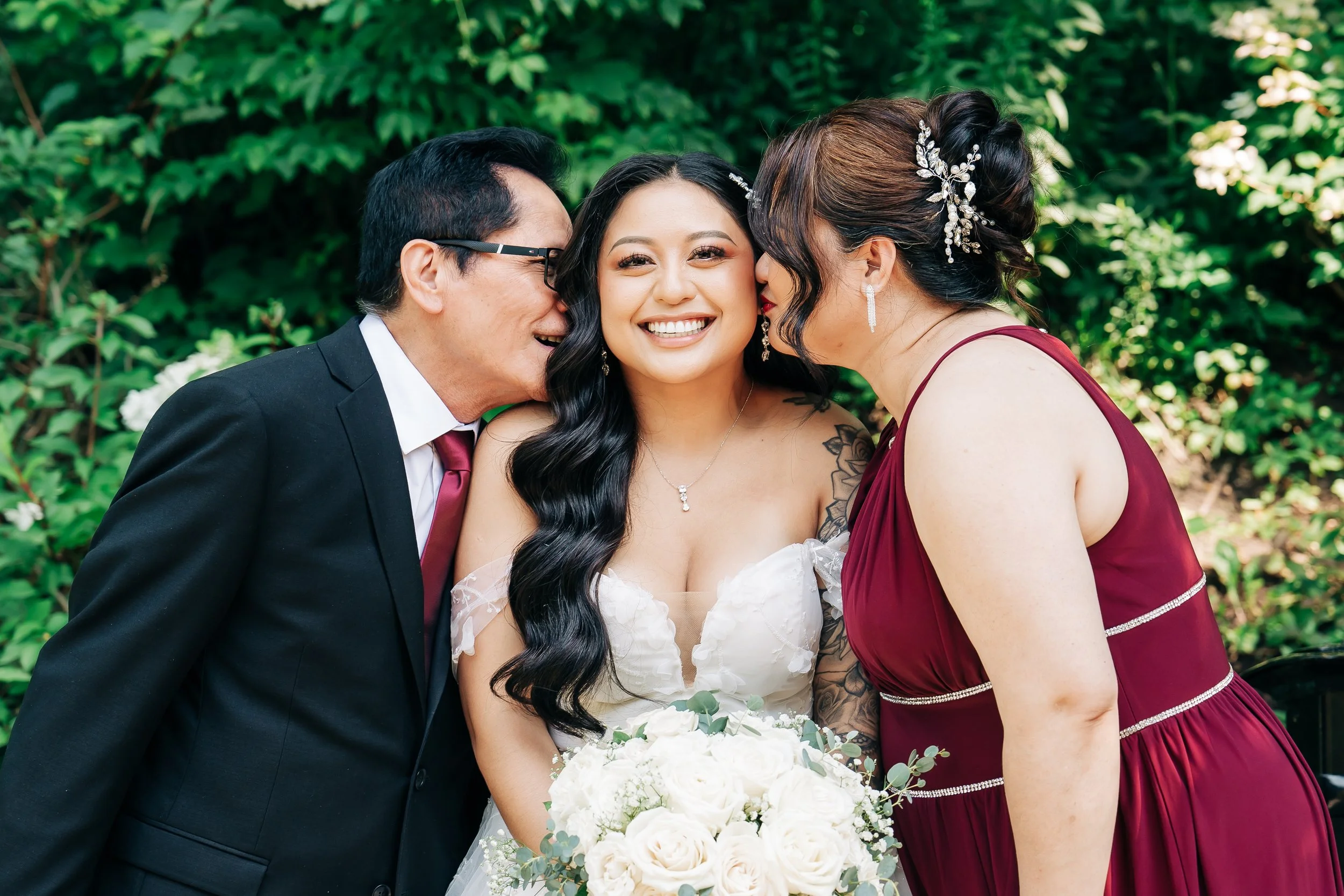 A bride with long black hair in a white wedding dress is being kissed on the cheek by her father in glasses and a man in a suit and tie, while a woman in a burgundy dress leans in to kiss her on the other cheek, all smiling amid green foliage.