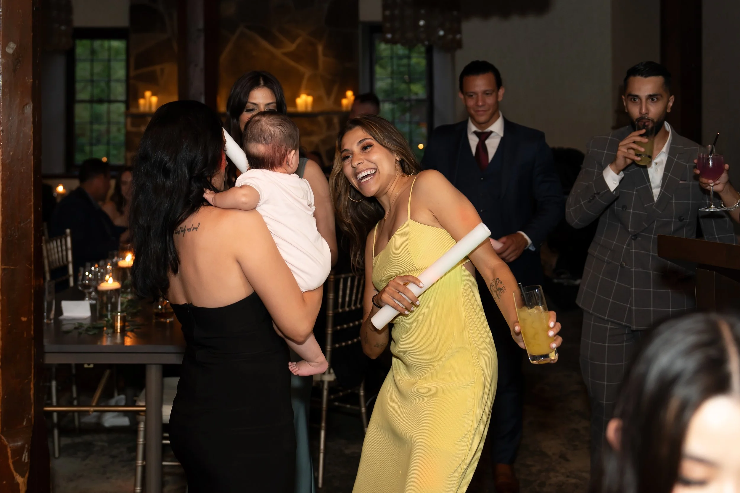 People celebrating at a party, with women holding a baby, one wearing a yellow dress, and others in formal attire, standing around a table with candles
