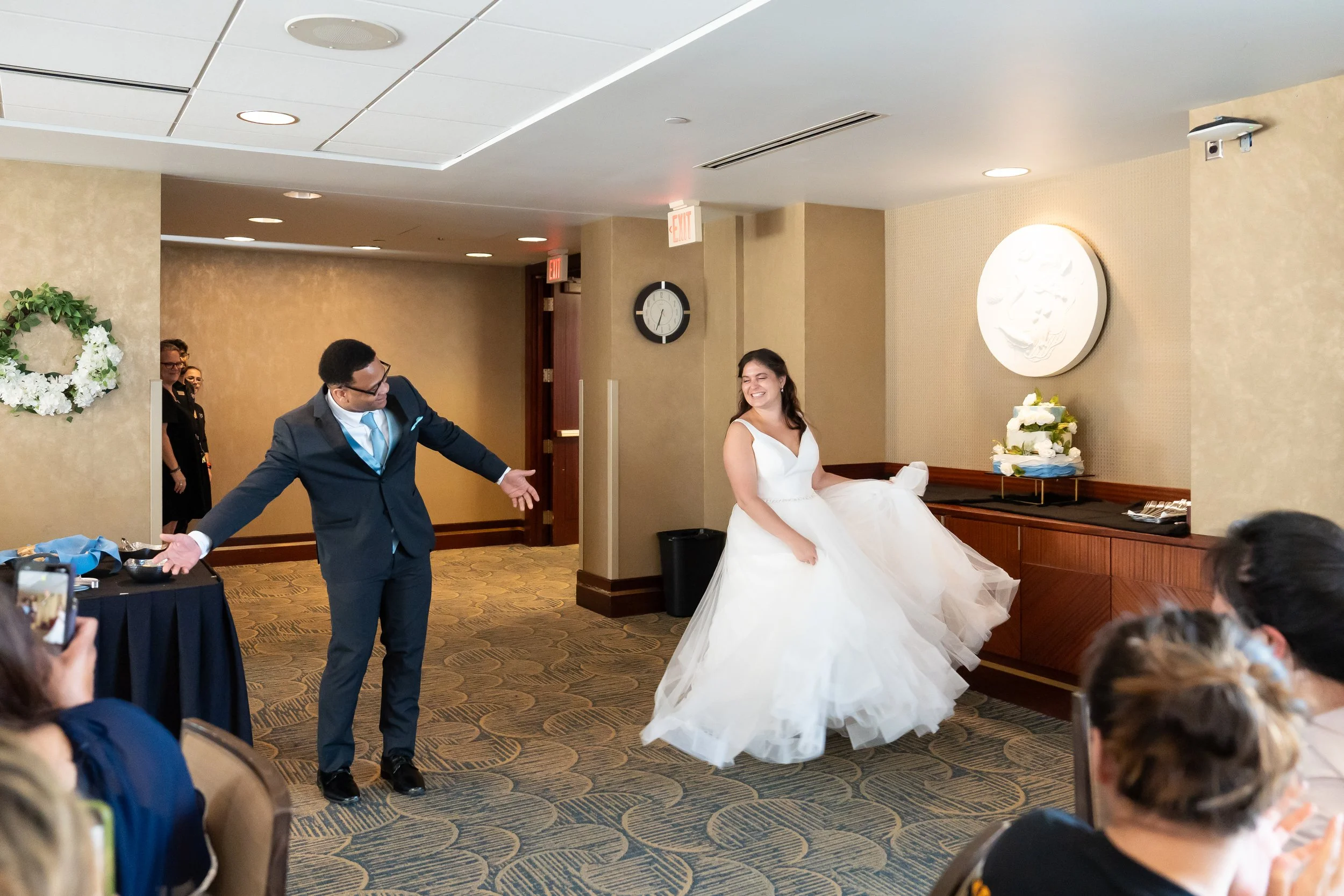 Bride in a wedding dress smiling and spinning, groom in a suit with arms wide open, wedding guests sitting and watching, indoor reception hall with decorated walls and a cake on a side table.