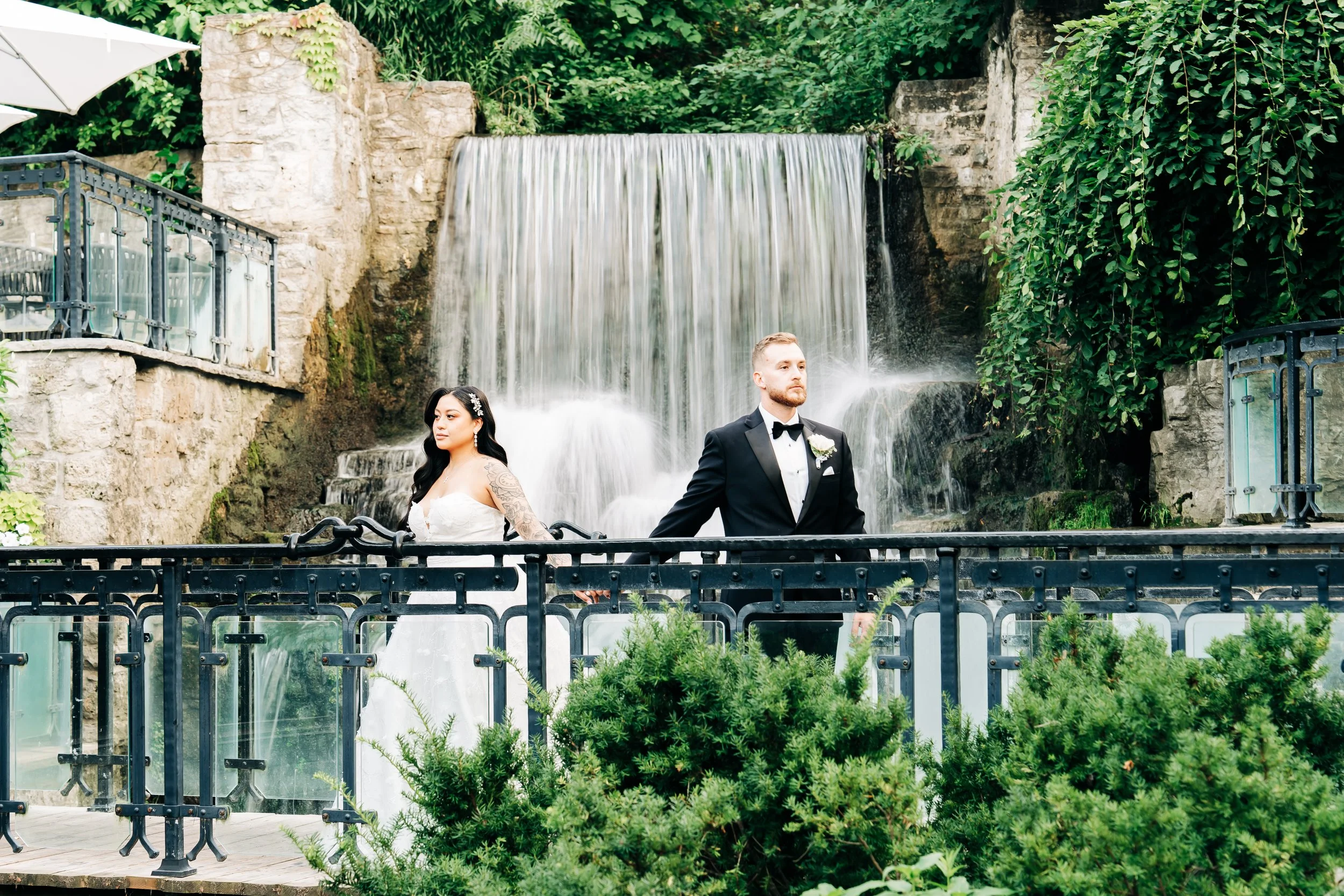 A bride and groom stand on a balcony in front of a waterfall, surrounded by greenery.
