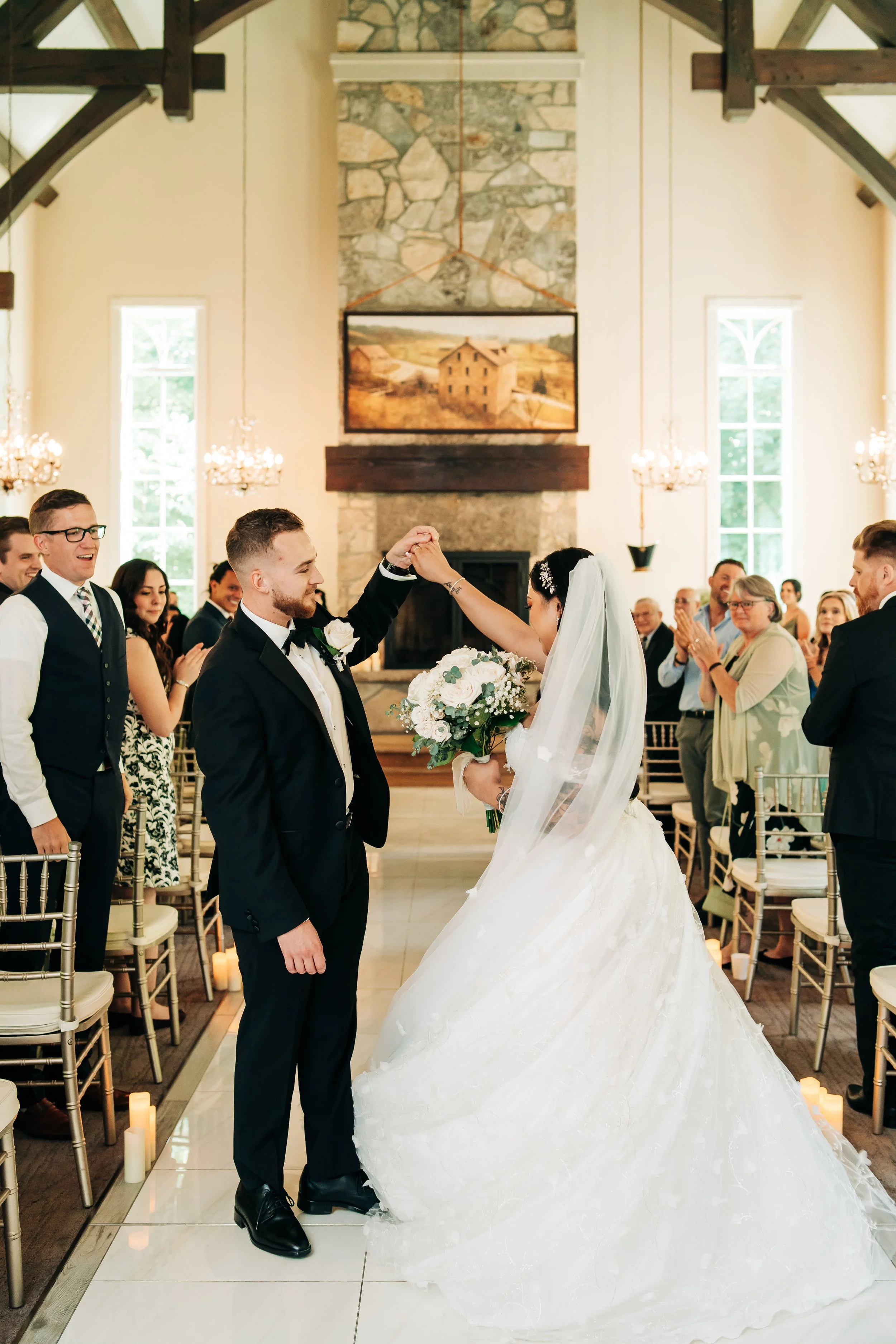 A bride and groom dancing during their wedding ceremony in a bright, elegant venue with guests clapping and smiling in the background.