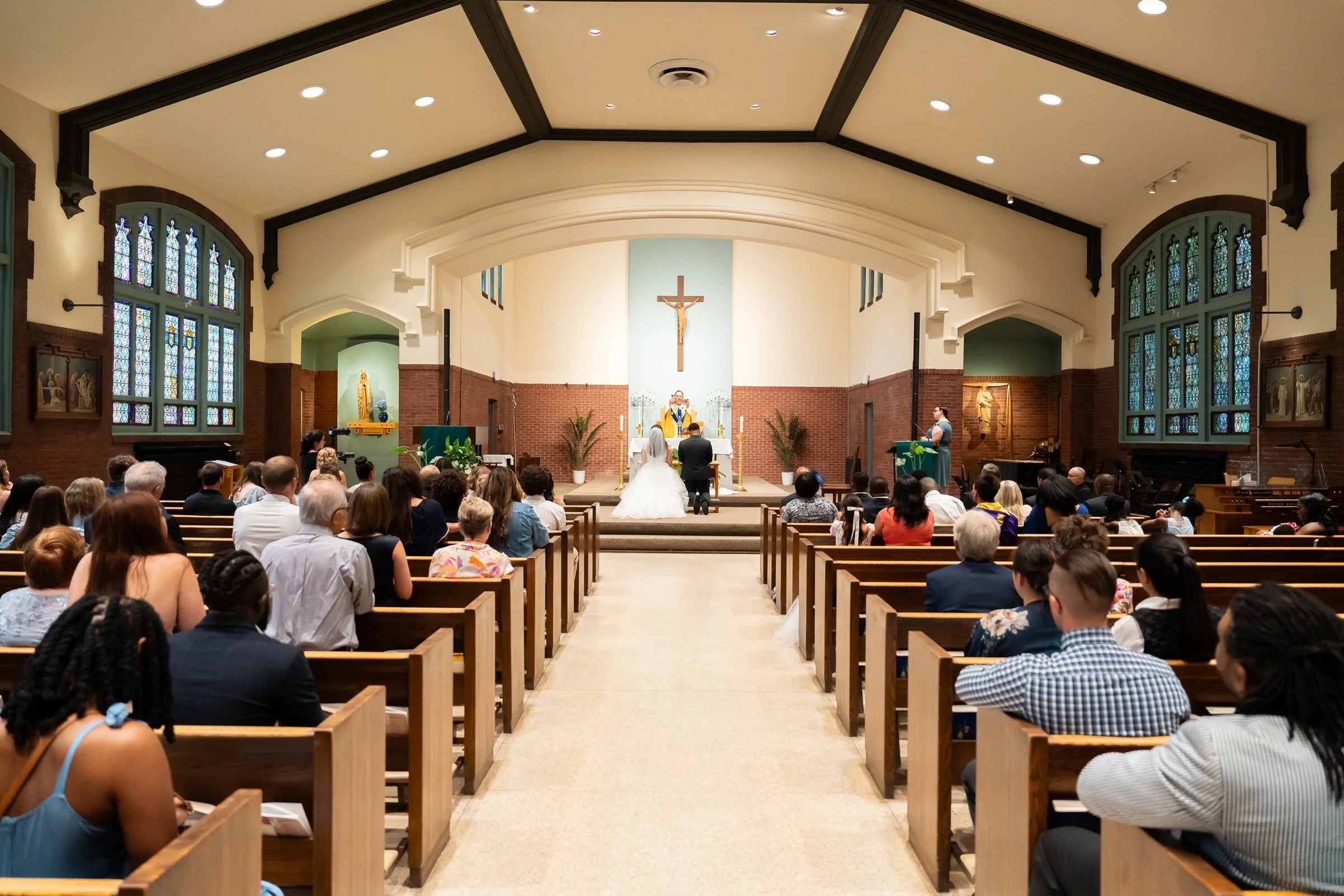 A wedding ceremony taking place inside a church with guests seated in pews facing the altar, where the couple is kneeling before the officiant.
