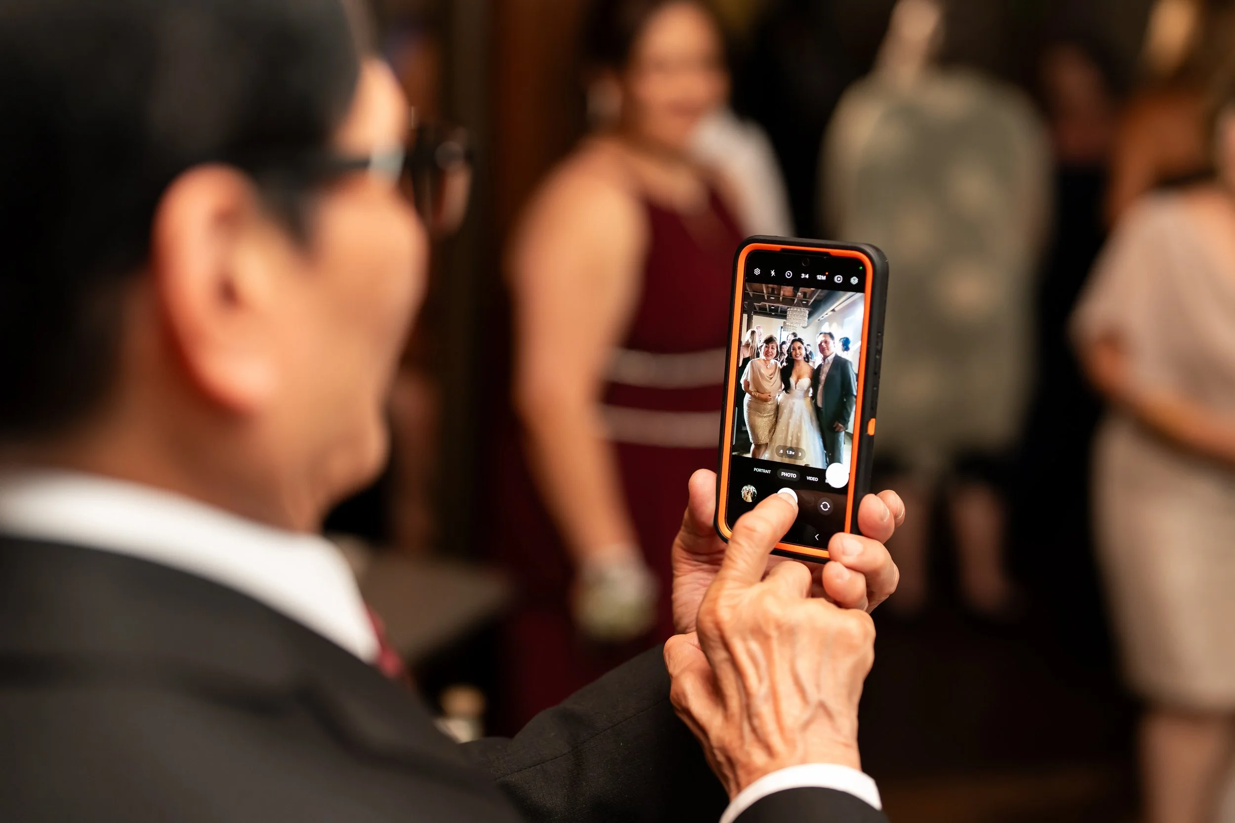 An elderly woman taking a photo with a smartphone at a formal event, with people in dresses and suits in the background.