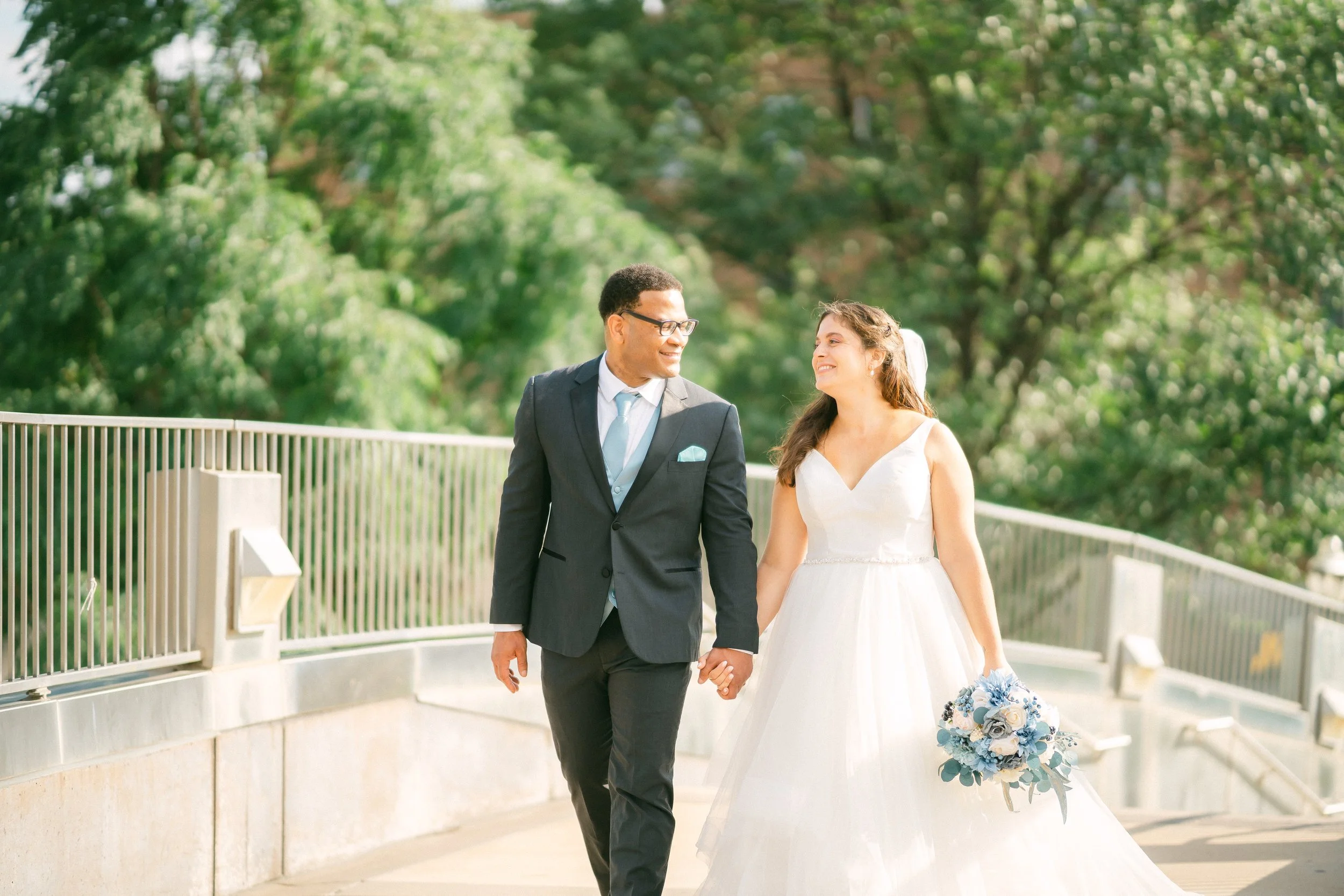 A bride and groom walking hand in hand outdoors during daytime, surrounded by green trees. The bride is in a white wedding dress holding a bouquet, and the groom is in a dark suit with glasses.