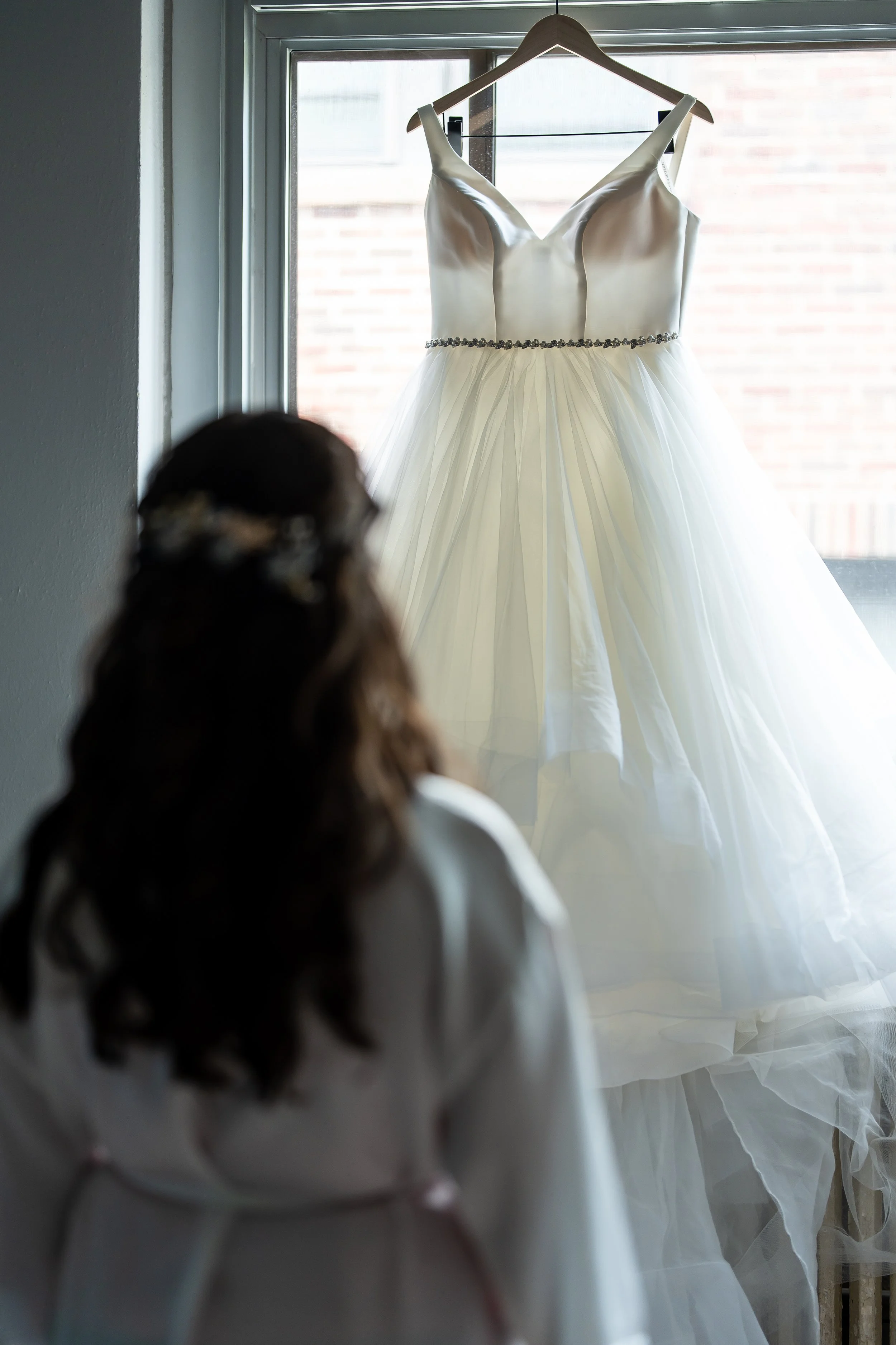 A woman with brown hair, seen from behind, watching a white wedding dress hanging on a hanger in front of a window.