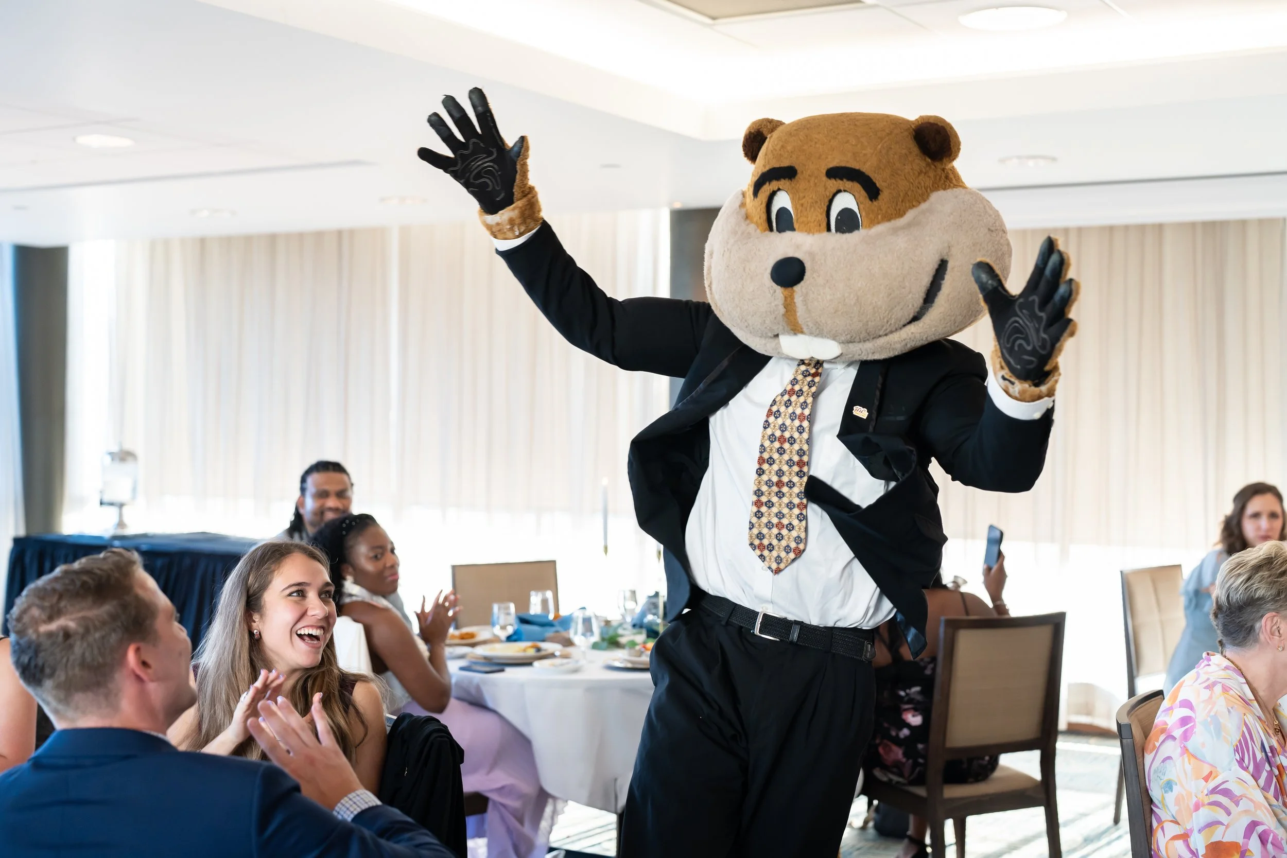 A person in a bear mascot costume, dressed in a suit and tie, entertaining guests at a banquet or conference table in a well-lit room.