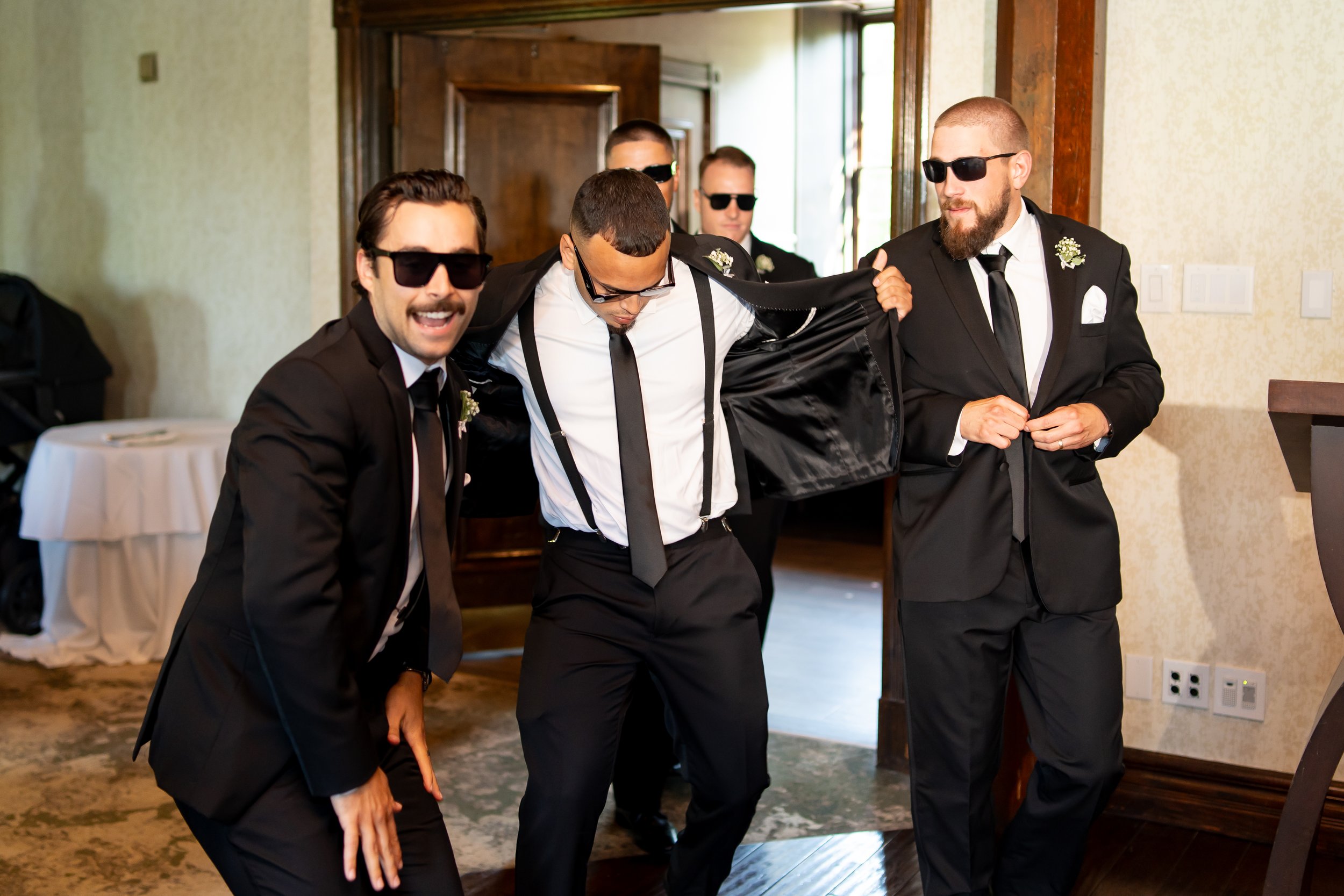 Group of men in black suits and sunglasses at a formal event, with one man opening his jacket as others smile and pose for the camera.
