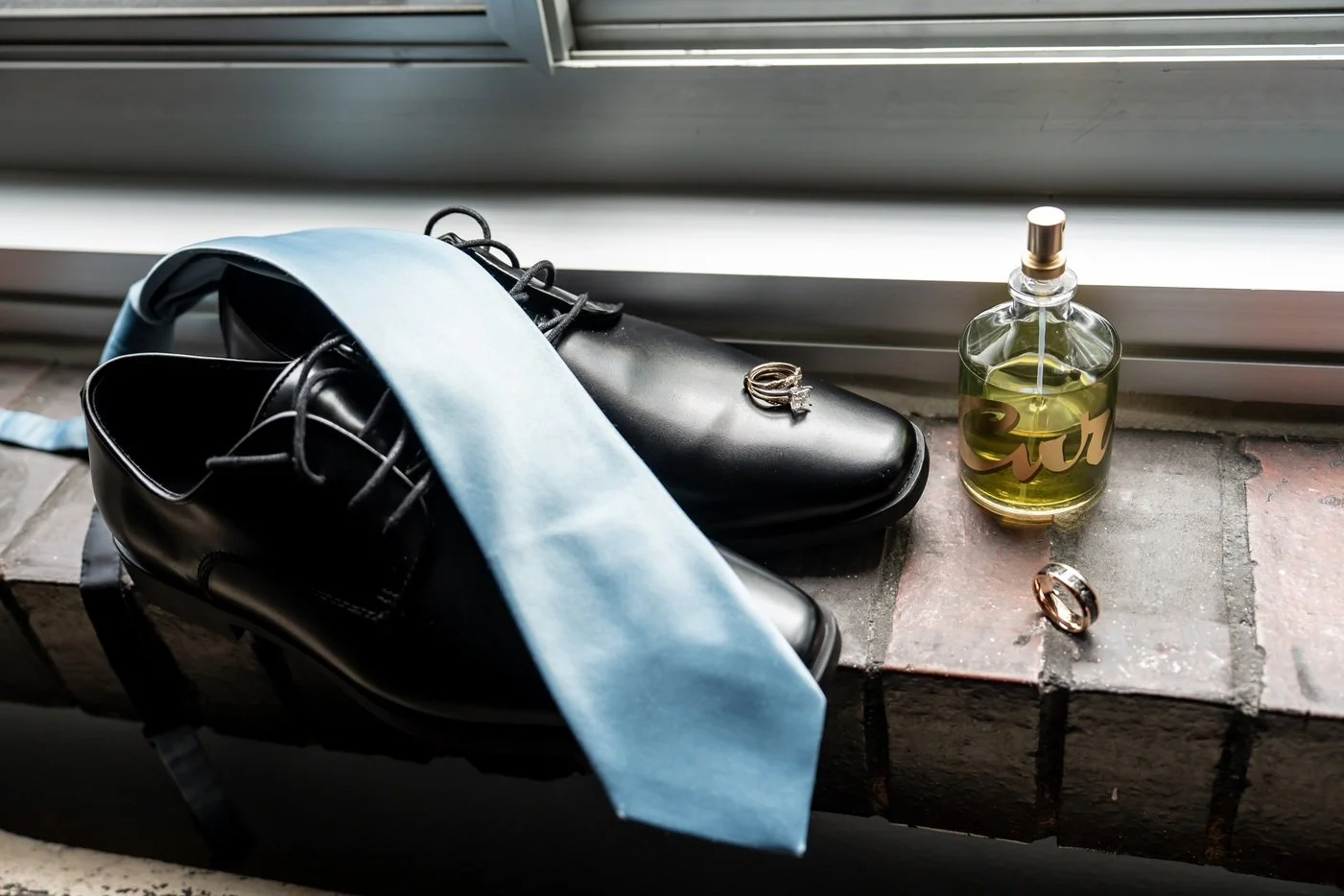 Black dress shoes with a silver ring resting on top, a light blue tie draped over the shoes, a bottle of perfume with a gold and clear design, and two rings, all placed on a brick window sill.