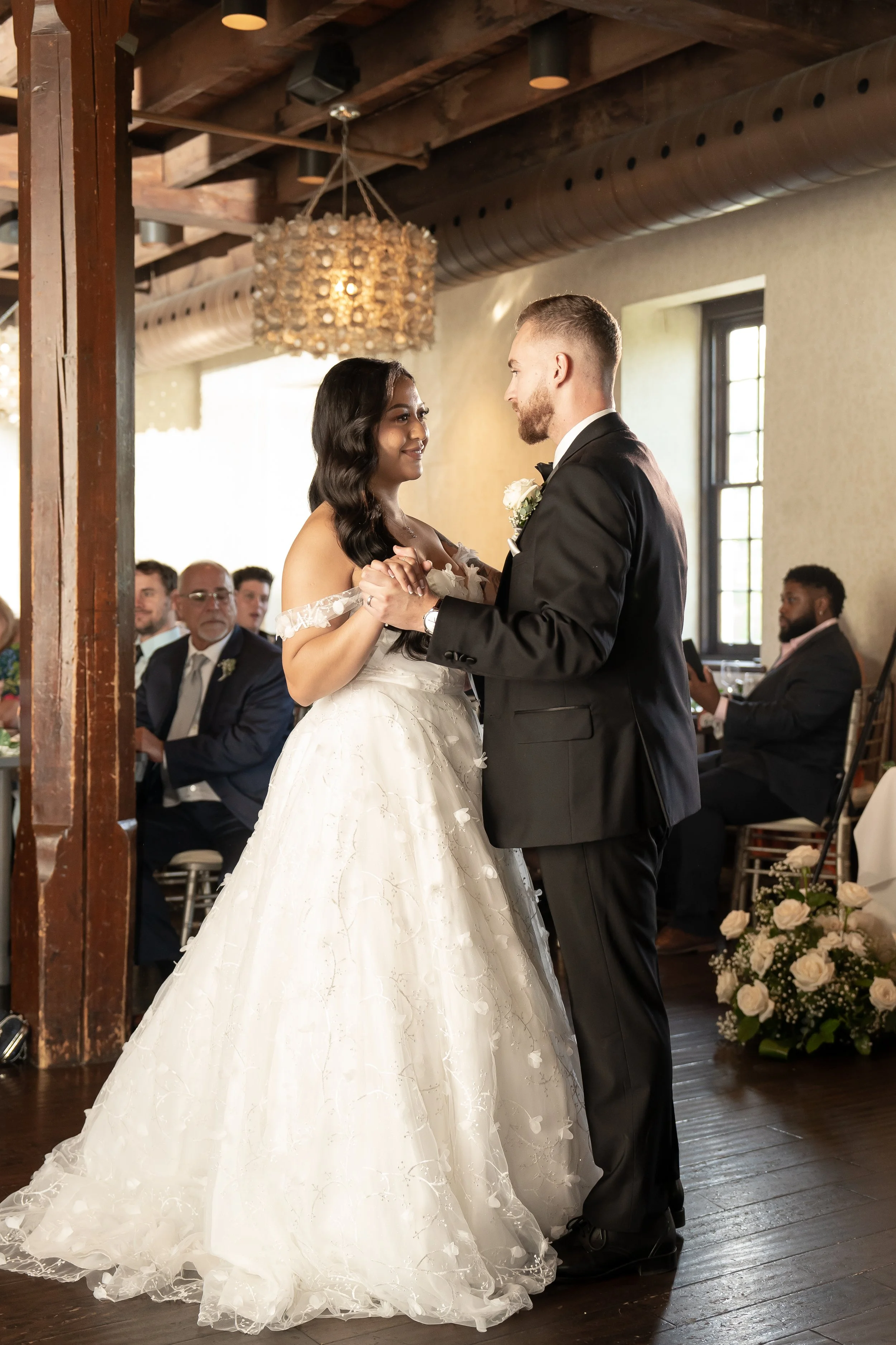A bride and groom dance during their wedding reception in a rustic venue with guests watching.