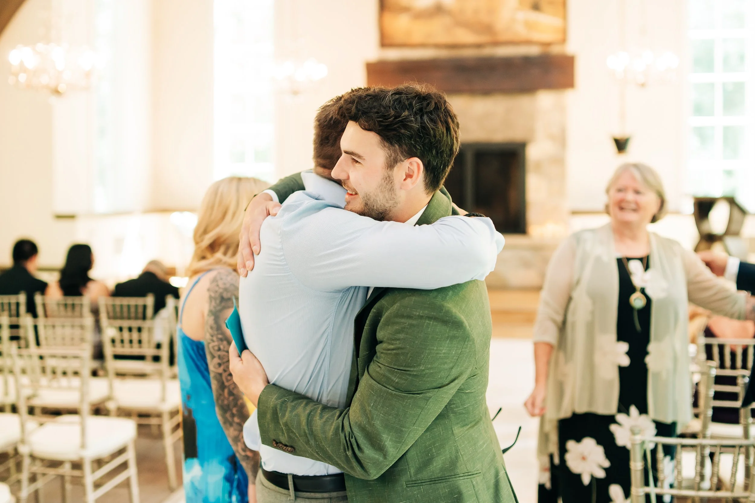 Two men hugging at a wedding reception, one with dark hair in a green blazer and the other with light hair in a light blue shirt, with guests and a fireplace in the background.