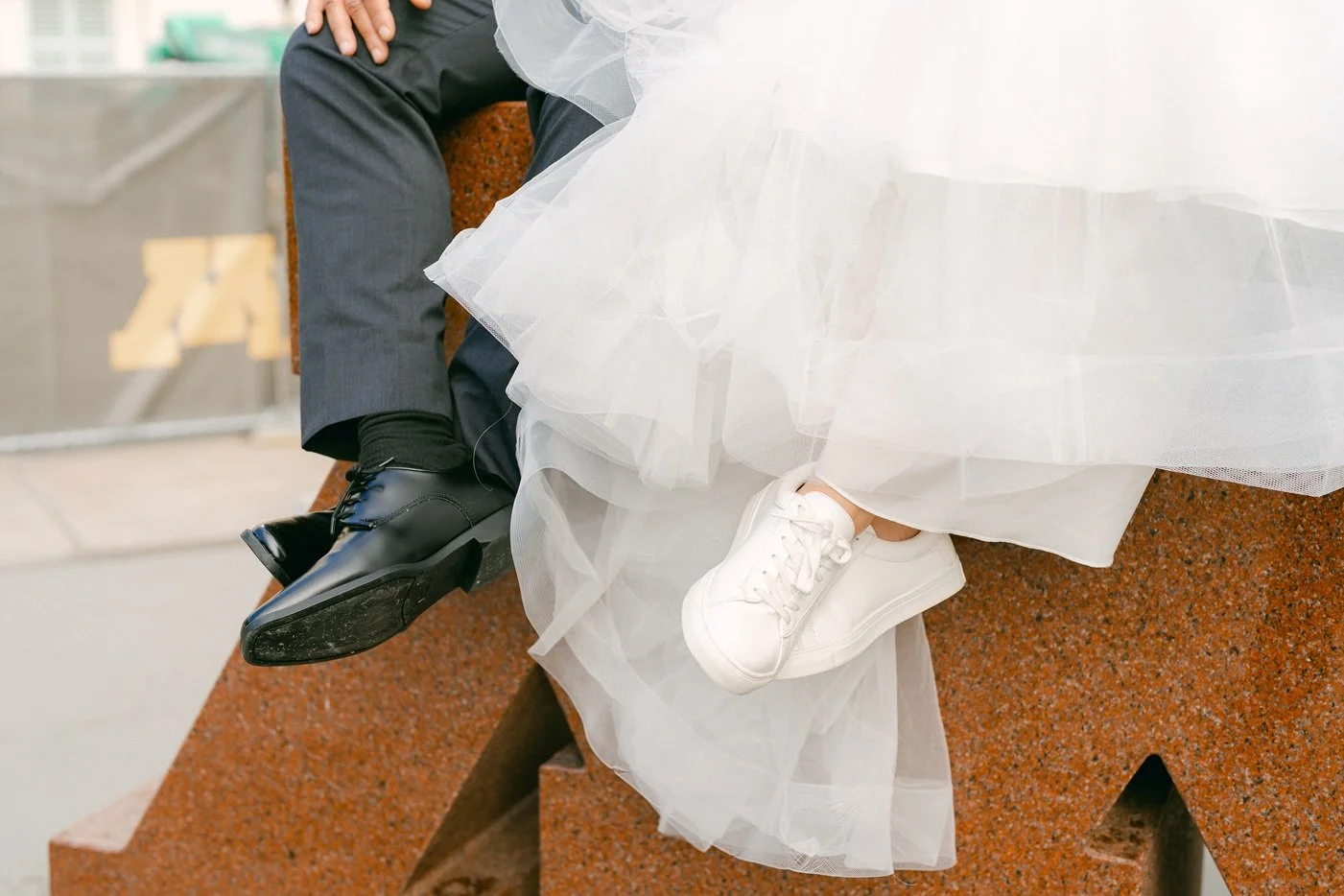Close-up of a couple sitting on a red stone bench, with only their legs visible. The woman is wearing white sneakers and a voluminous white dress, while the man is wearing black shoes and dark gray pants.