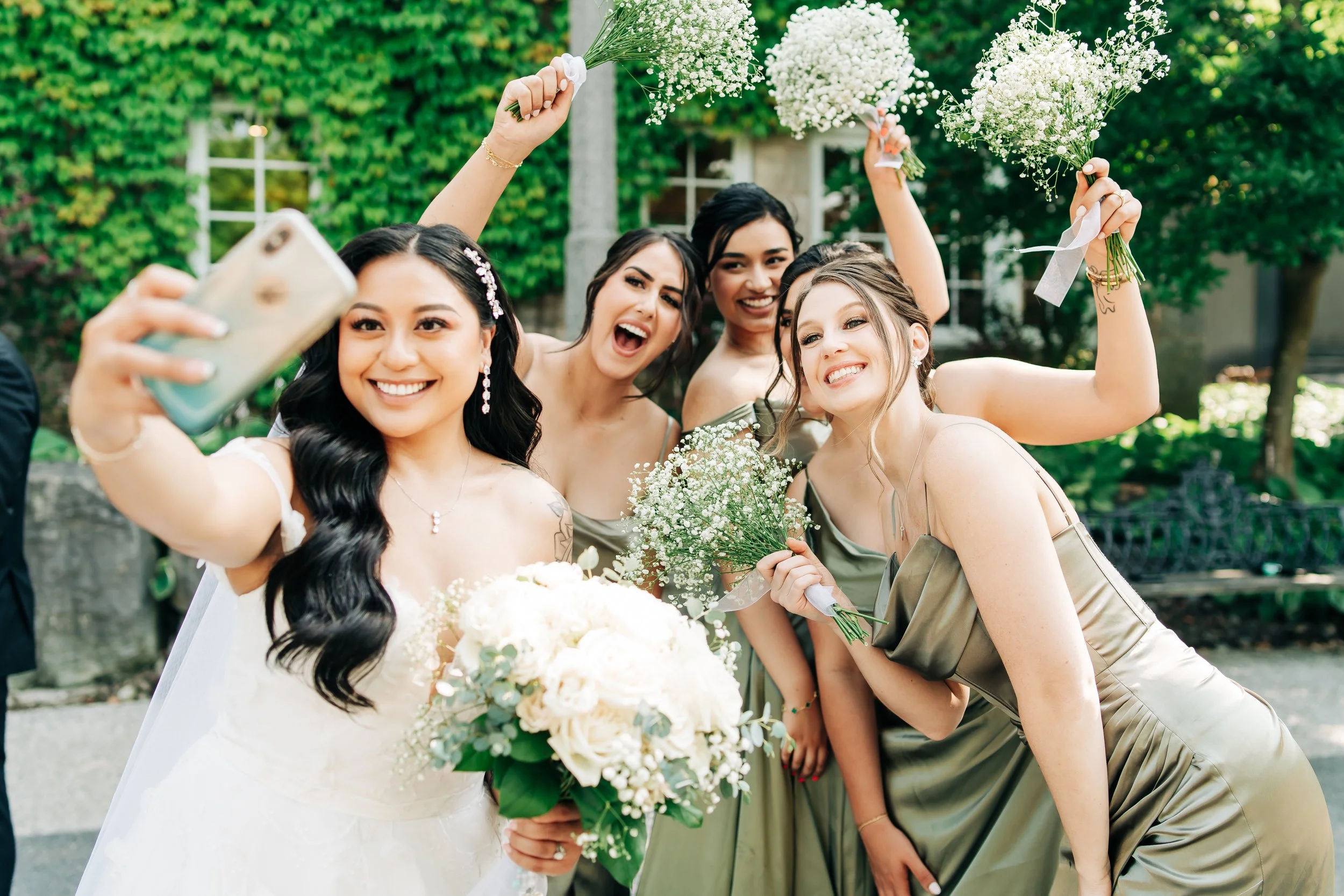 A bride in a white wedding dress is taking a selfie with four bridesmaids in green dresses, all holding bouquets of white flowers outdoors.