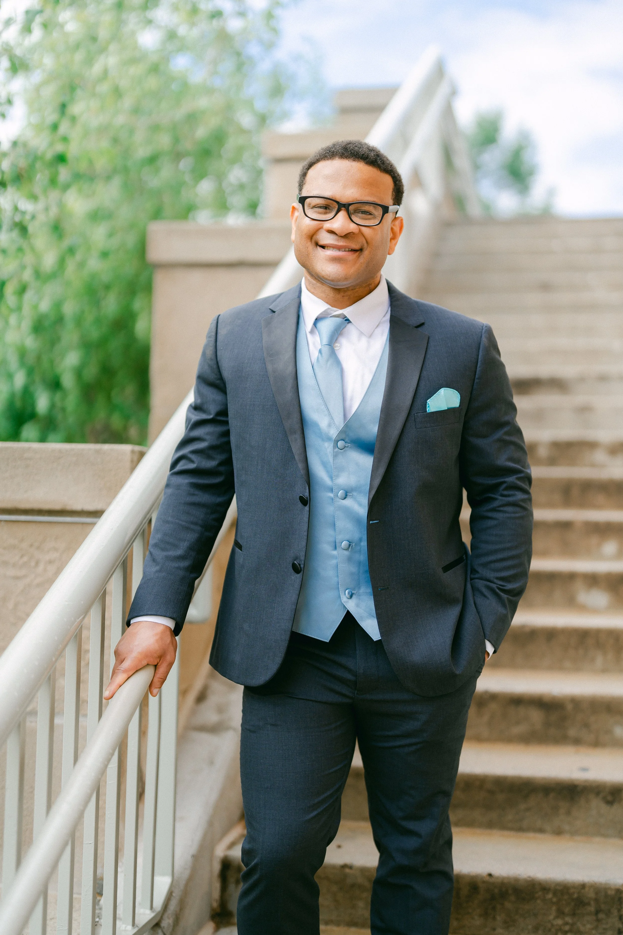 Smiling man in a dark blue suit with a light blue vest and tie standing on outdoor stairs.