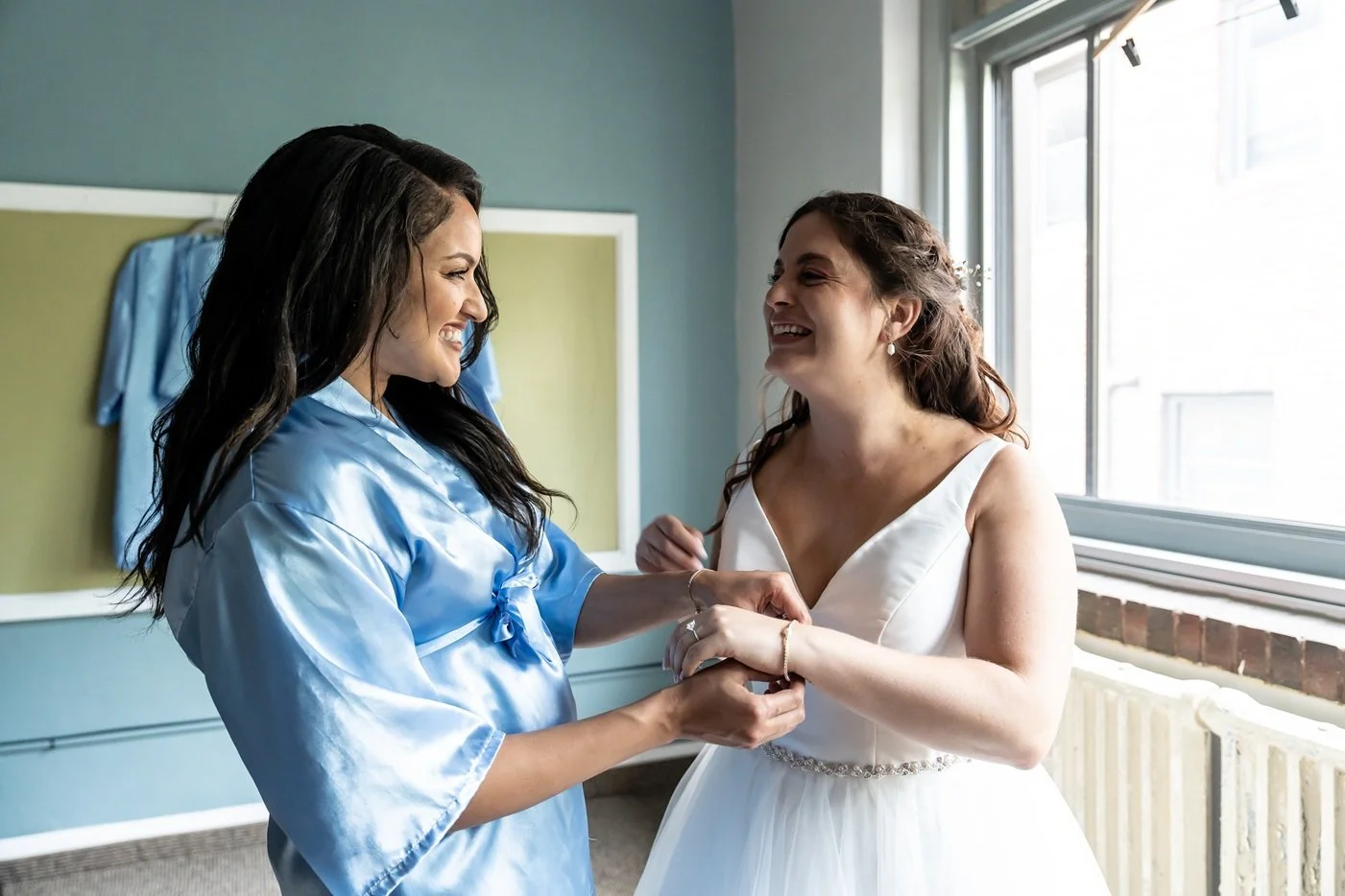 A bride and her friend sharing a joyful moment as the friend helps her into her wedding dress in a well-lit room with large windows.