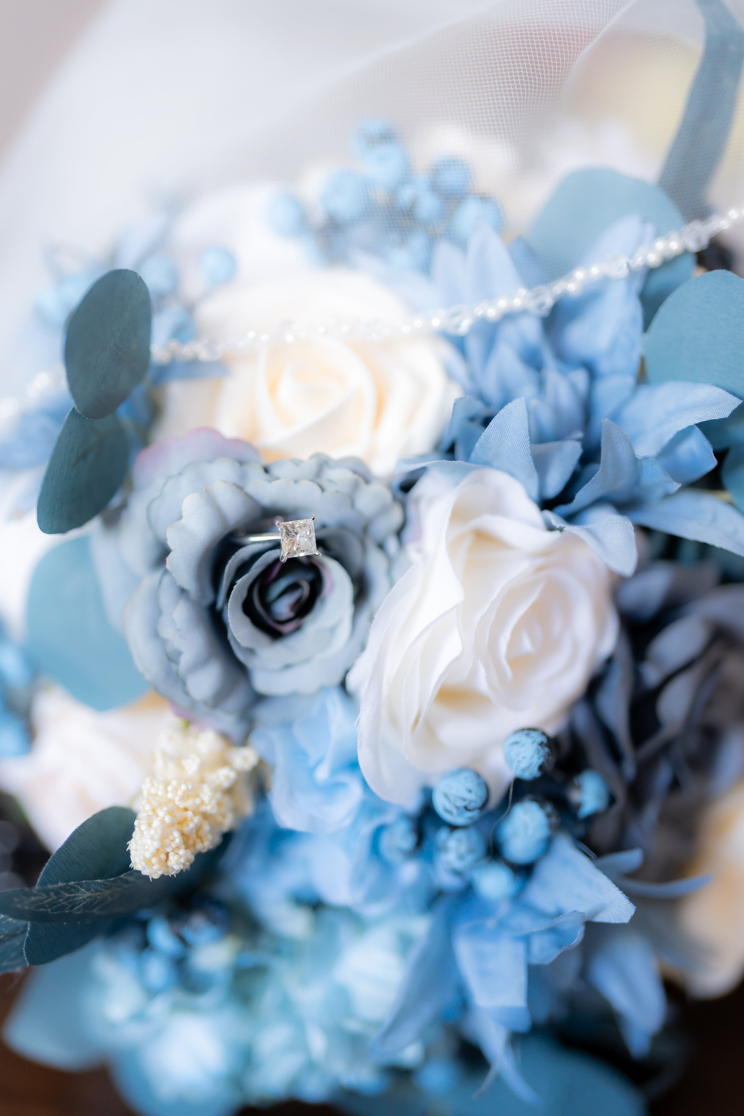 Close-up of a wedding bouquet with white and blue flowers, a pearl necklace, and an engagement ring with a square diamond.