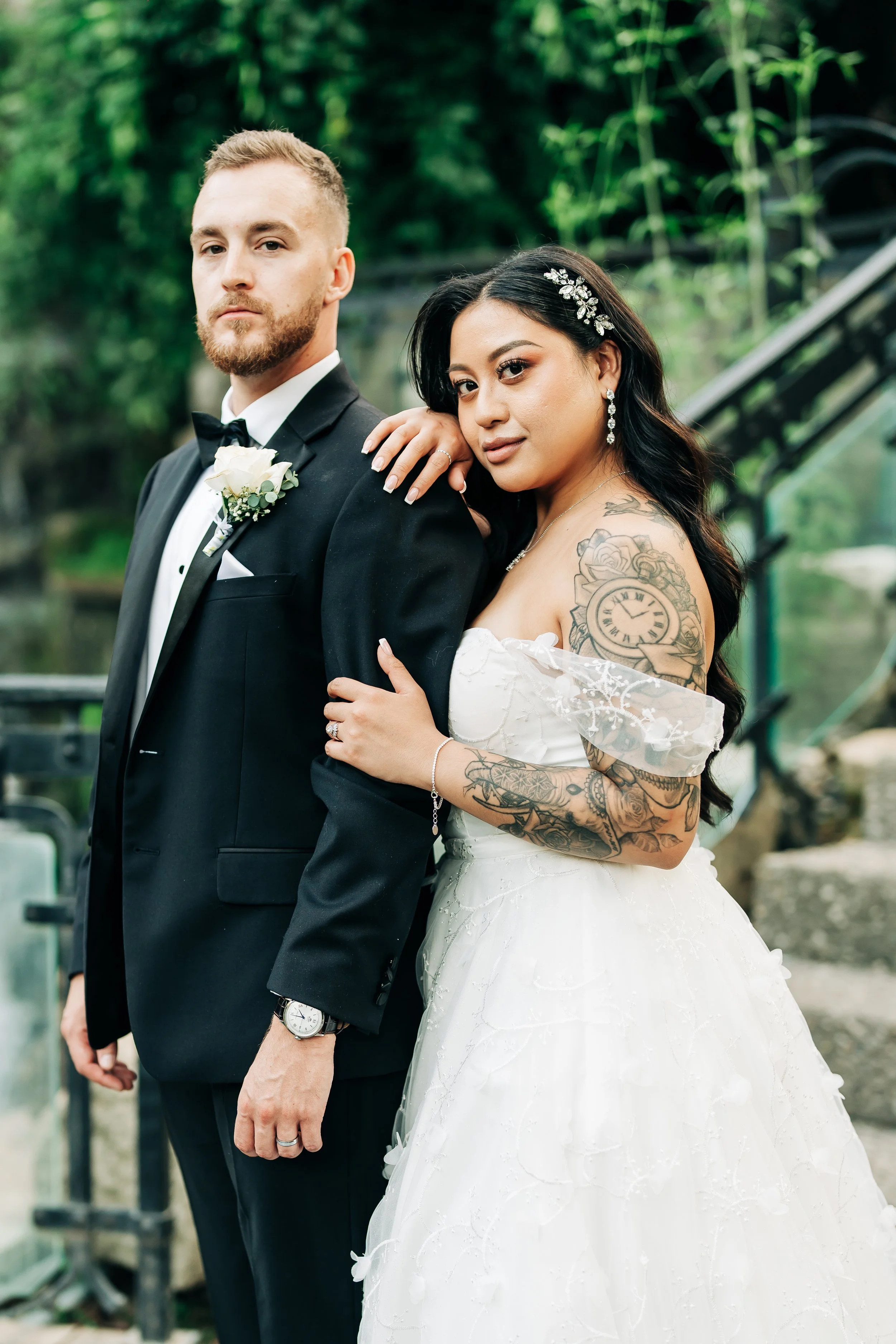 A bride and groom posing outdoors, the bride has tattoos and is dressed in a white wedding gown, the groom is in a black tuxedo with a white boutonniere.