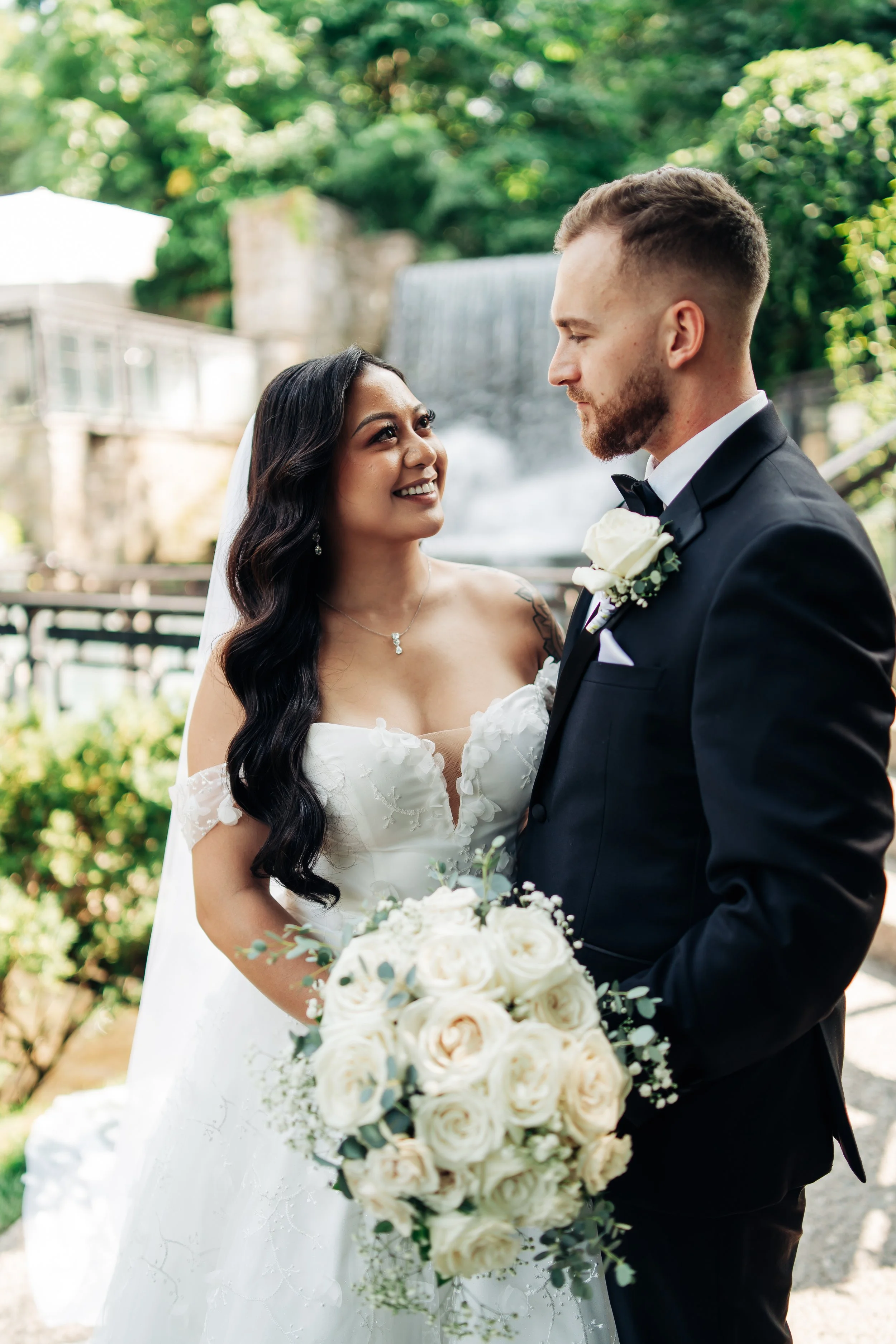 Bride and groom gazing at each other outdoors, with greenery and a waterfall in the background. The bride wears a white wedding dress and holds a bouquet of white roses, while the groom is dressed in a black suit with a white boutonniere.