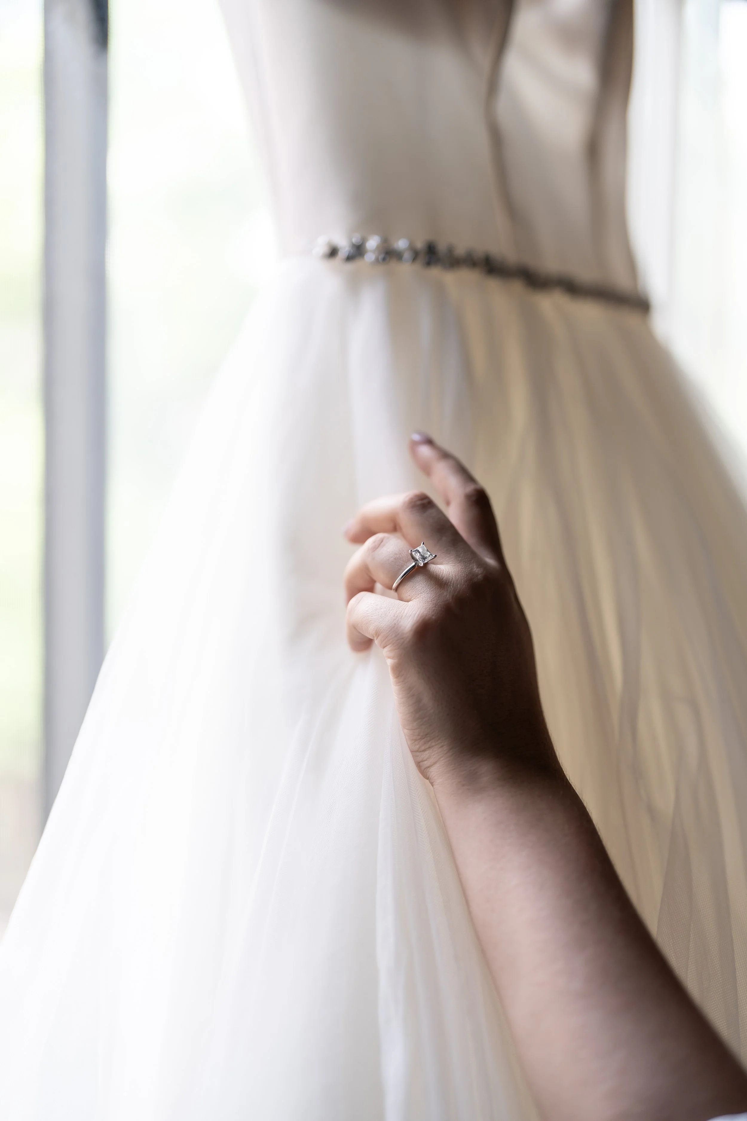 Close-up of a woman's hand with a wedding ring, holding the fabric of a wedding dress, near a window.
