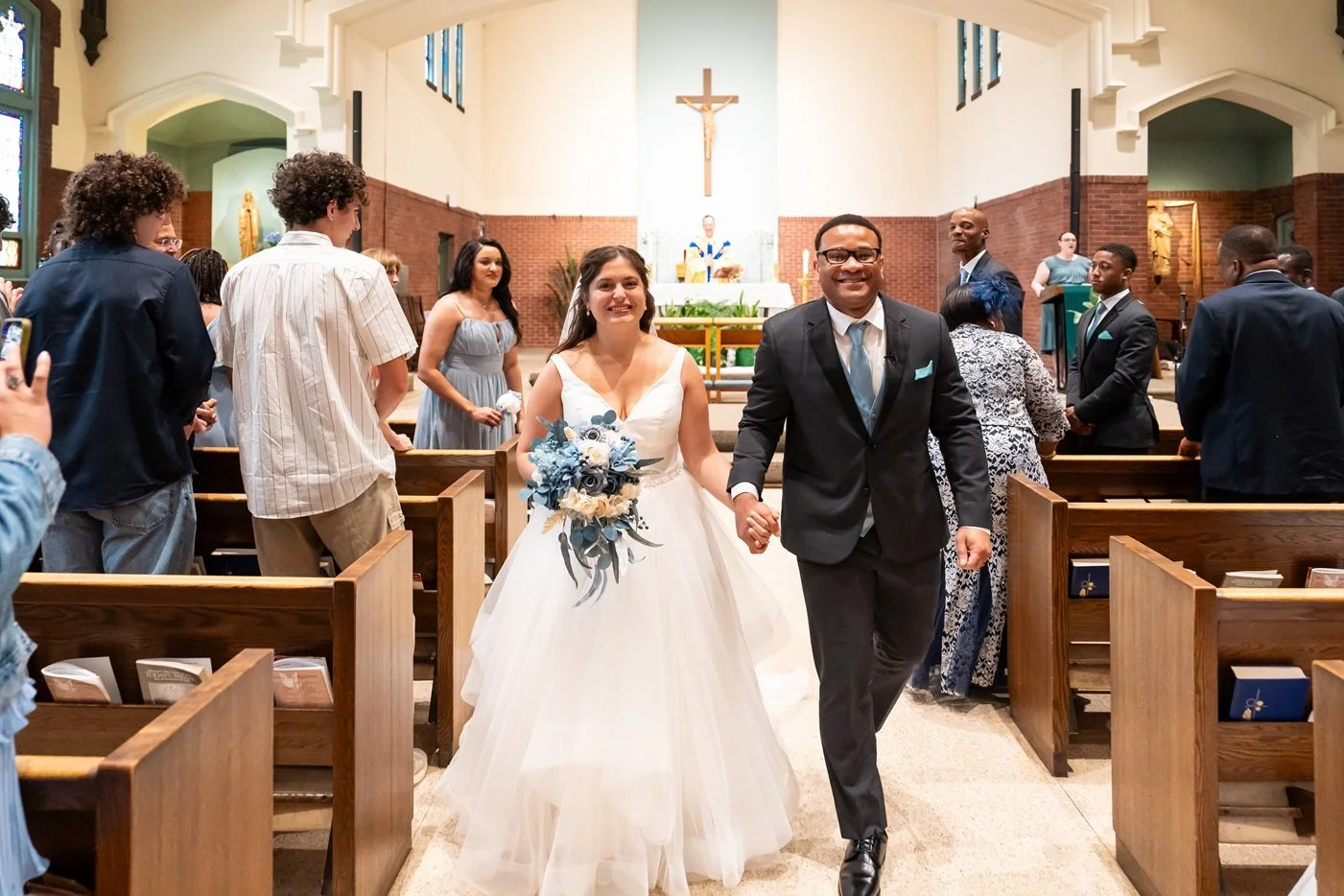 Bride and groom walking hand in hand down the aisle after their wedding ceremony inside a church, with guests seated and standing on either side.