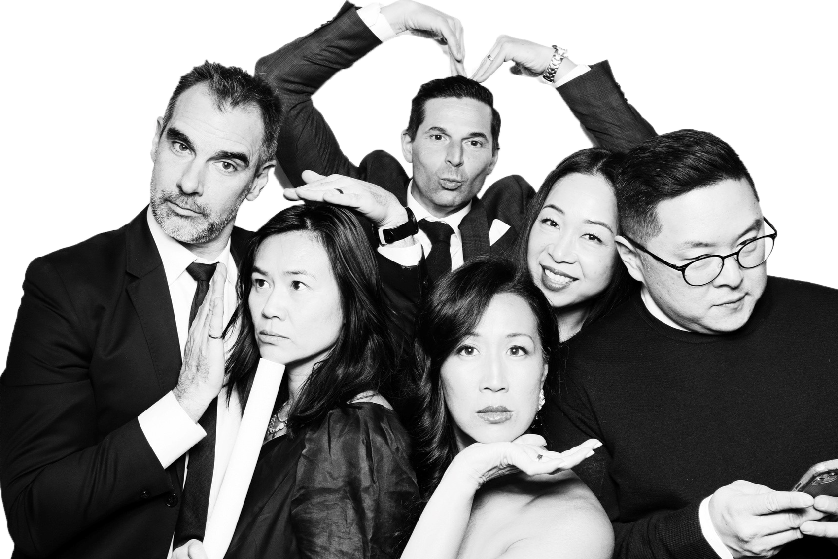 Group of six diverse adults in formal attire posing playfully for a black and white photo, with some making expressive gestures.
