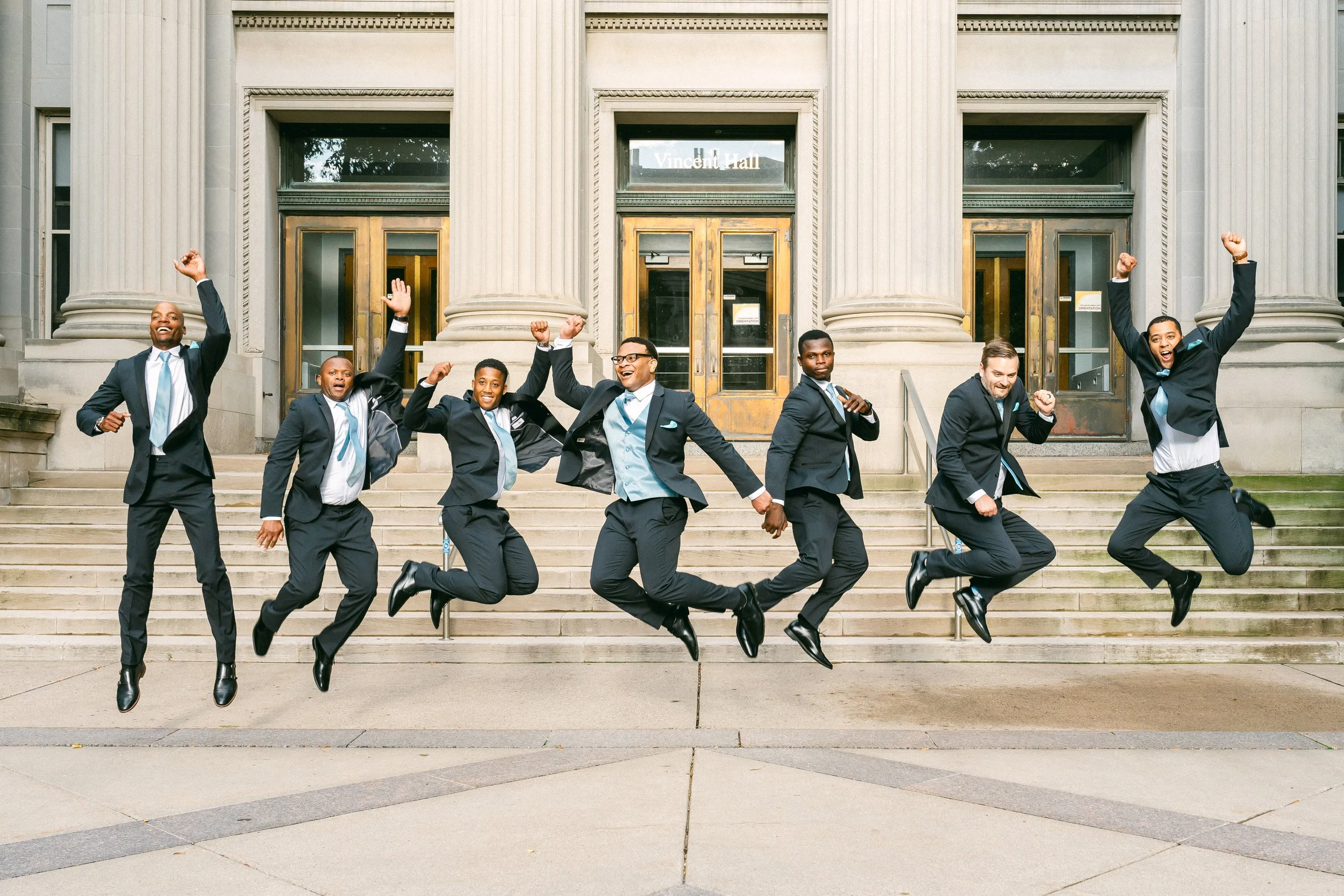 Seven men in suits jumping in front of a historic building with columns and stairs, celebrating.
