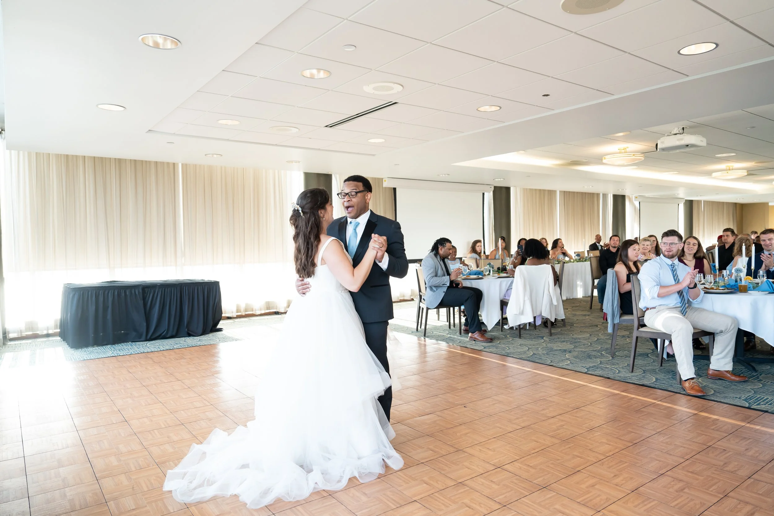 A bride and groom dance together at their wedding reception with guests seated at tables in the background.