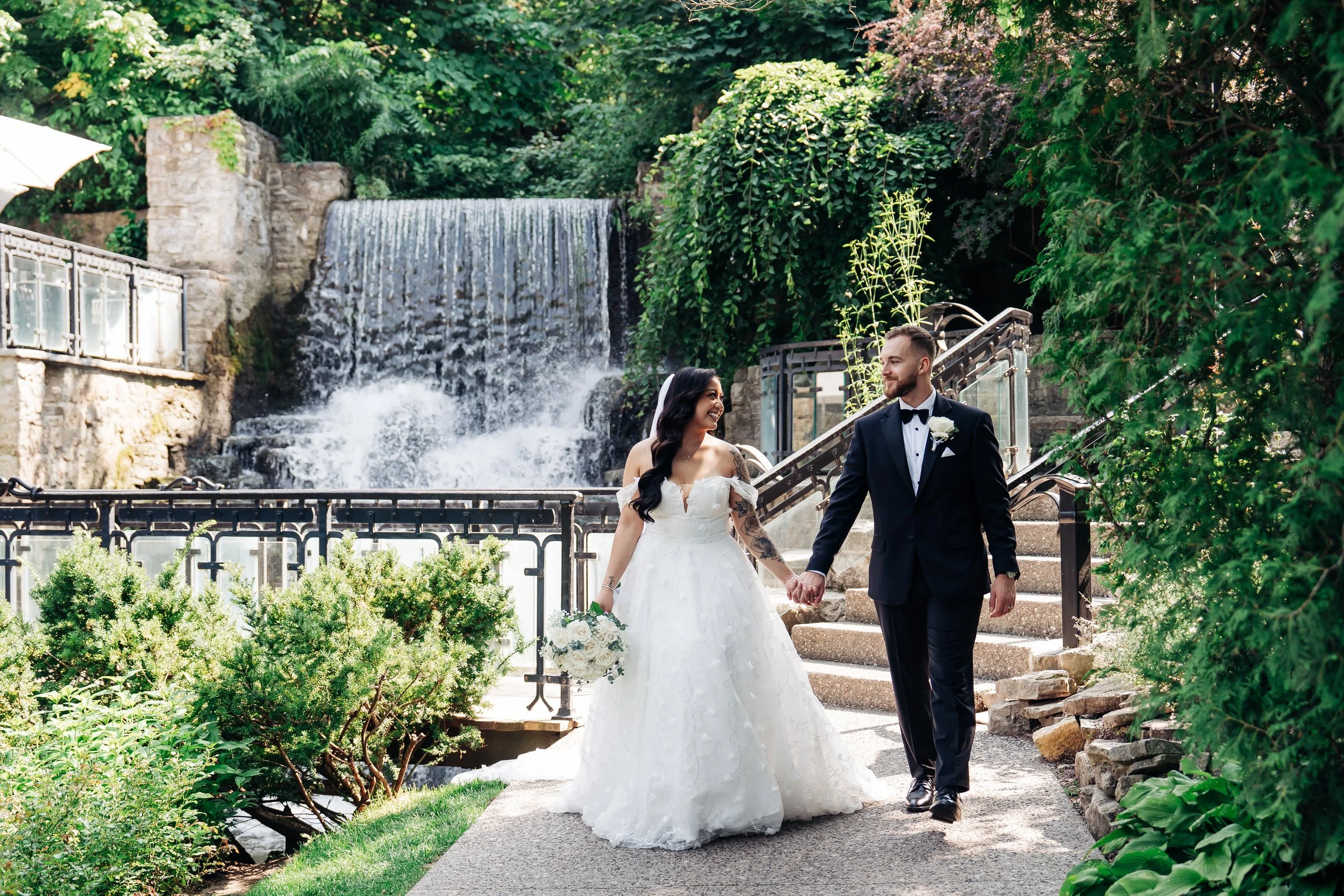 Bride and groom holding hands and walking together in a lush garden with a waterfall in the background.