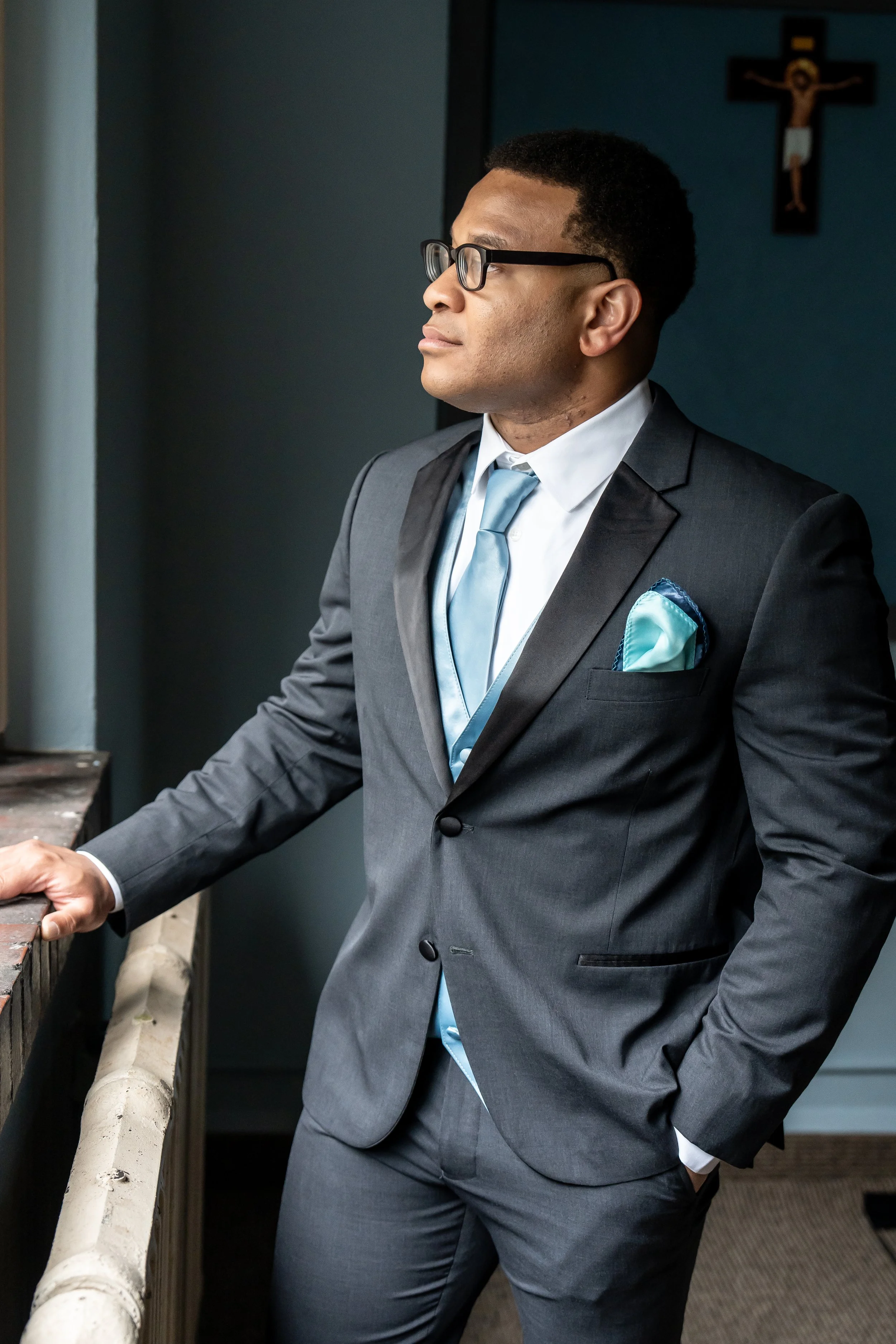 A man in a gray suit and light blue tie standing by a window, looking out with a contemplative expression, near a crucifix on the wall.