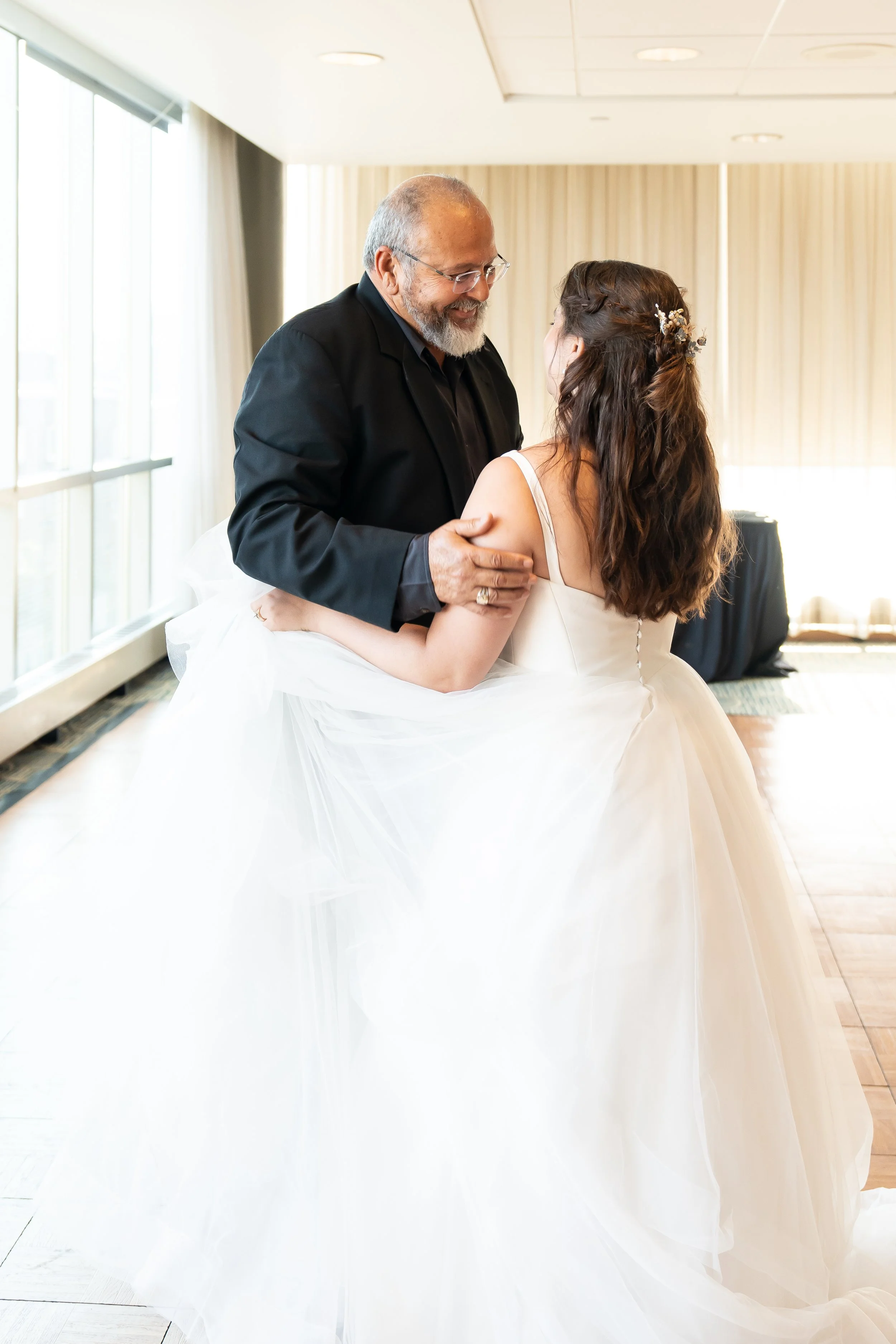 A bride and an older man sharing a dance, smiling at each other in a bright room with large windows and curtains.