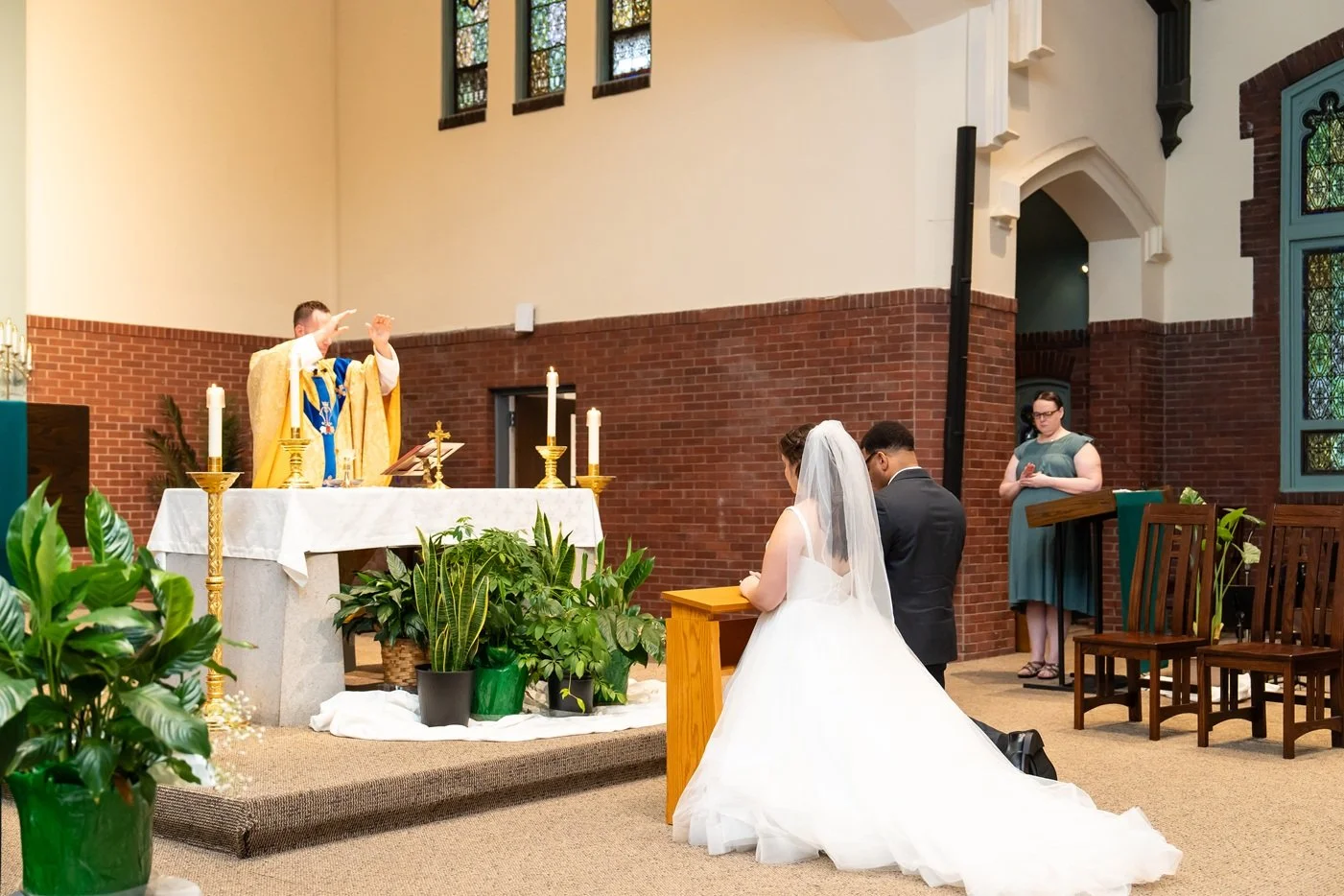 A bride and groom kneeling in prayer during a church wedding ceremony, with a priest and a deacon conducting the service, surrounded by green plants and candles.