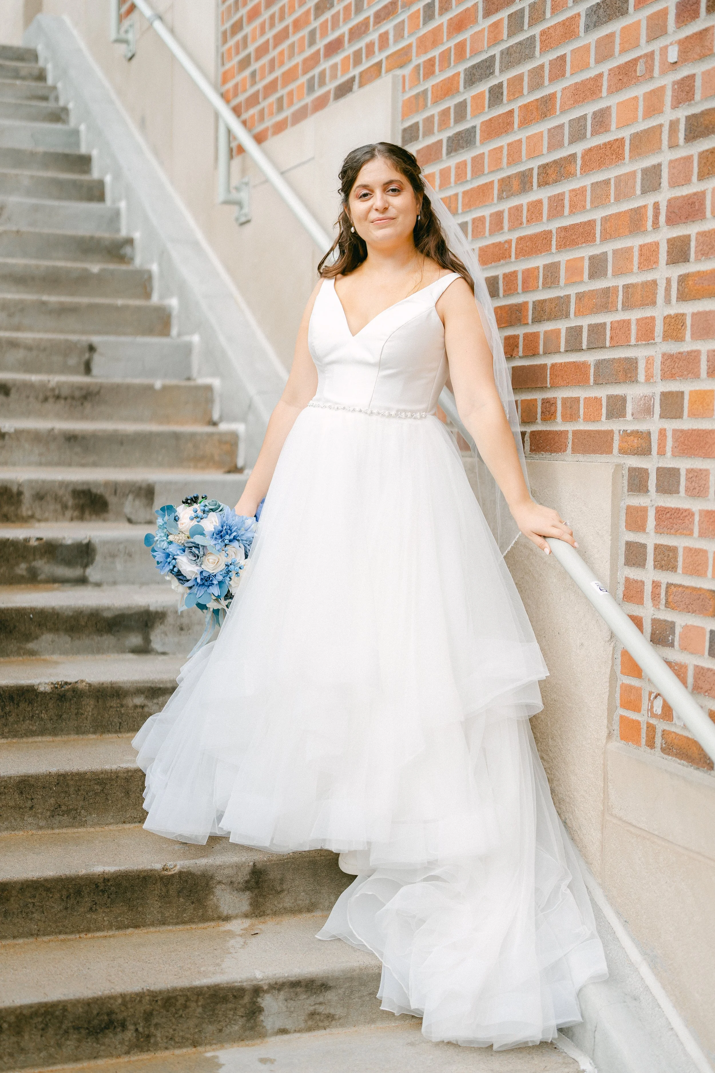 Bride in a white wedding dress holding a blue floral bouquet, standing on outdoor concrete stairs next to a brick wall.
