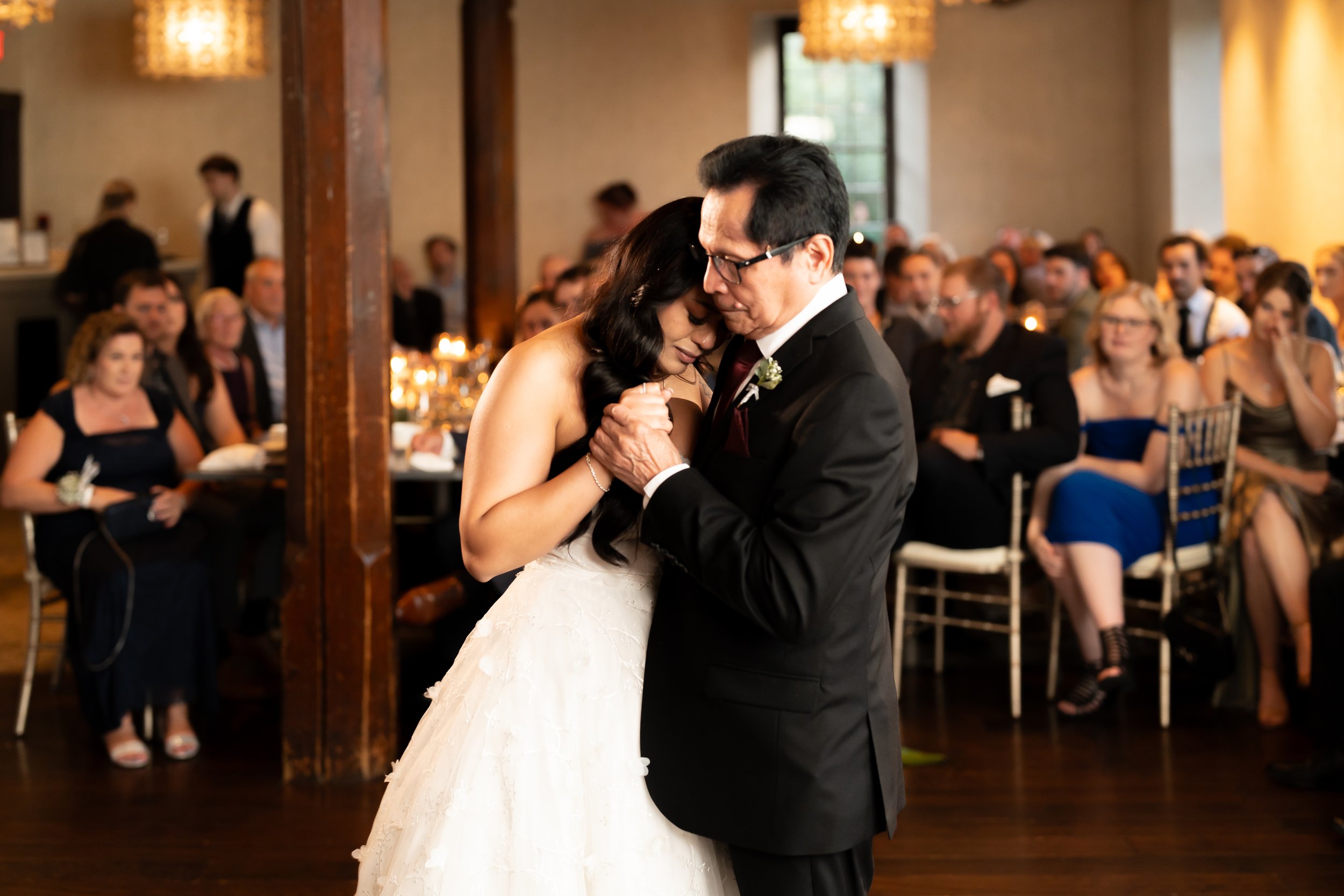 A bride and an older man, likely her father, sharing a dance at her wedding reception. The bride is wearing a white wedding gown, and the man is in a black suit. Guests are seated in the background watching them.