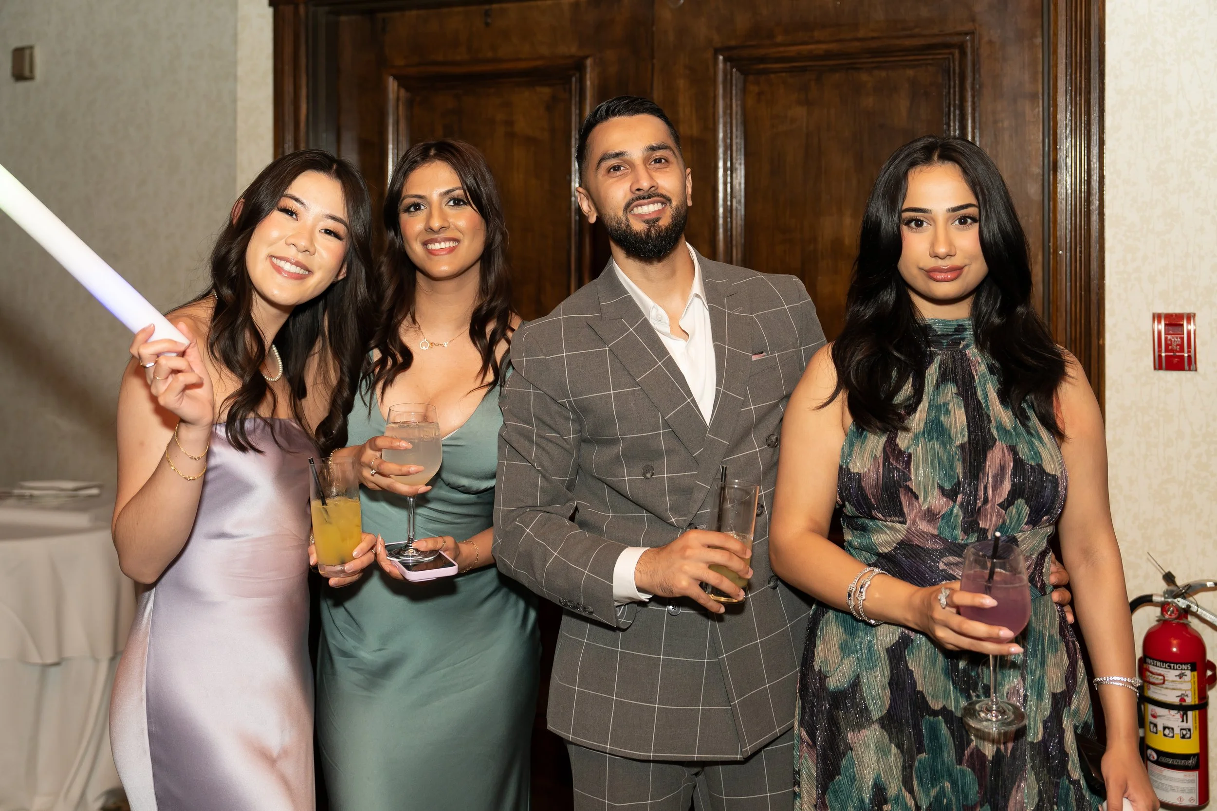 Four friends in evening attire at a party, smiling and holding drinks, standing in front of a wooden wall.