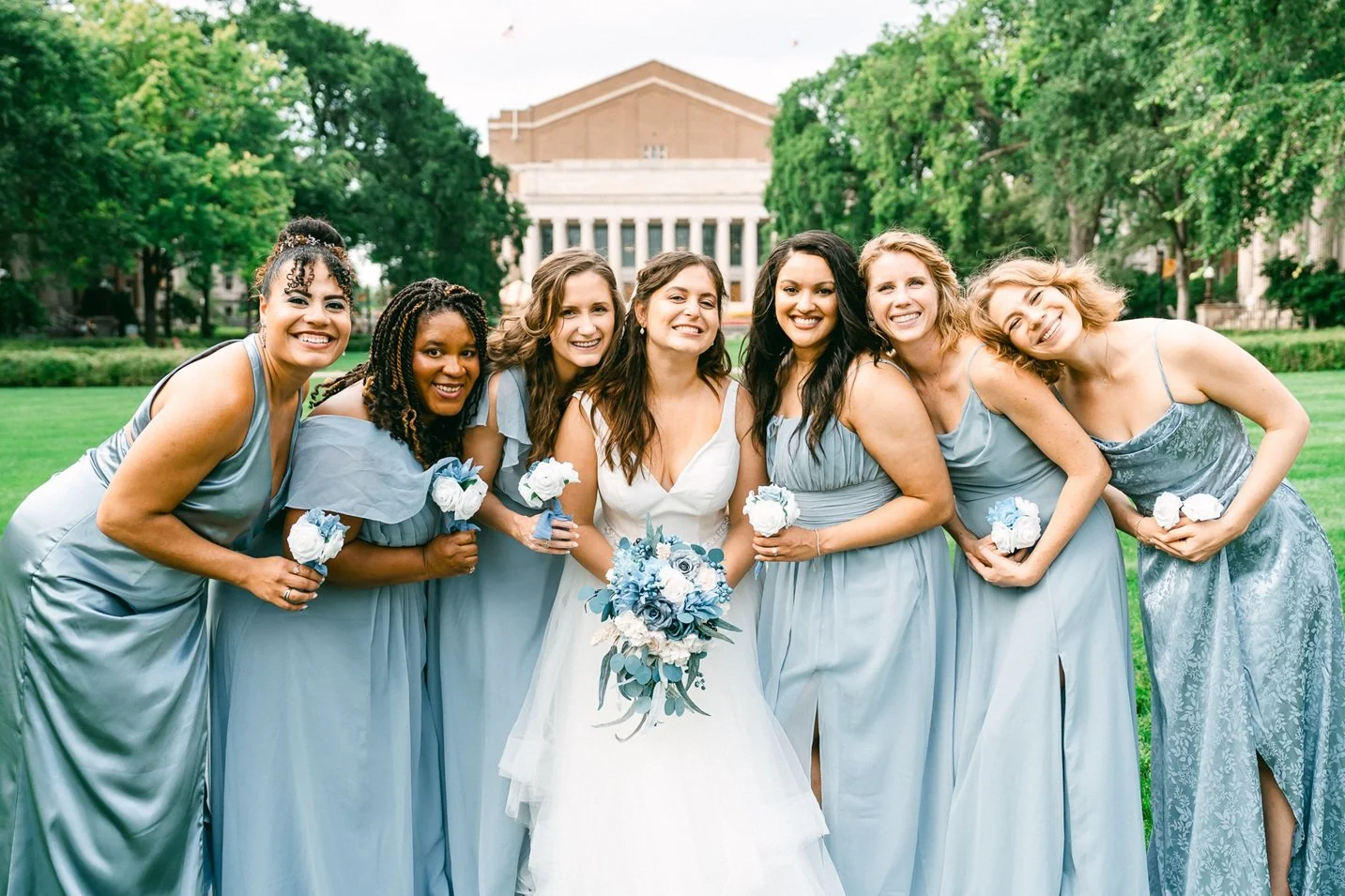 A bride wearing a white wedding dress holding a bouquet of blue and white flowers, standing with six bridesmaids in light blue dresses, each holding a white boutonniere, on a grassy lawn with trees and a building in the background.