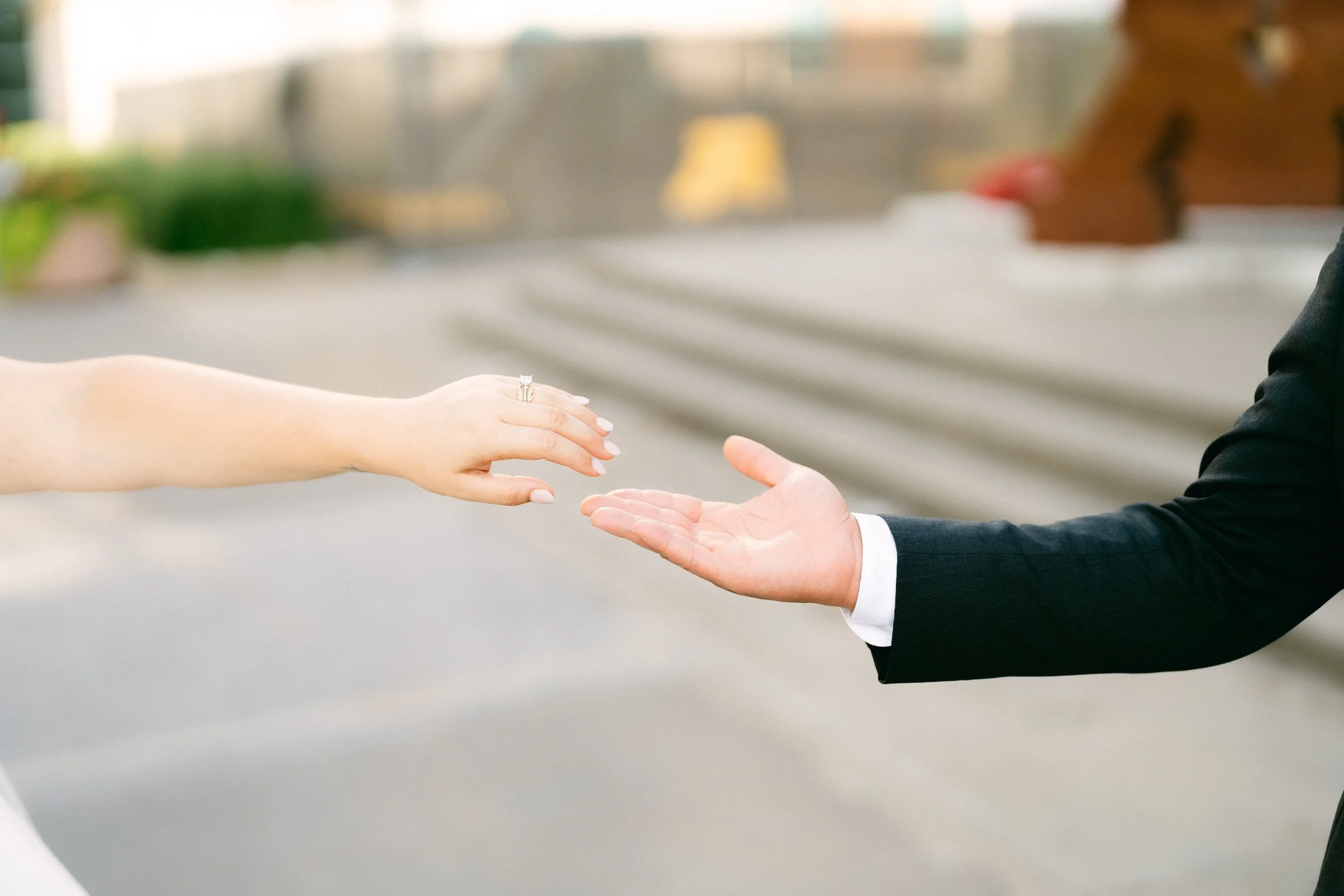 A woman in a wedding dress reaching out her hand towards a man in a suit with an open hand, against a blurred outdoor background.
