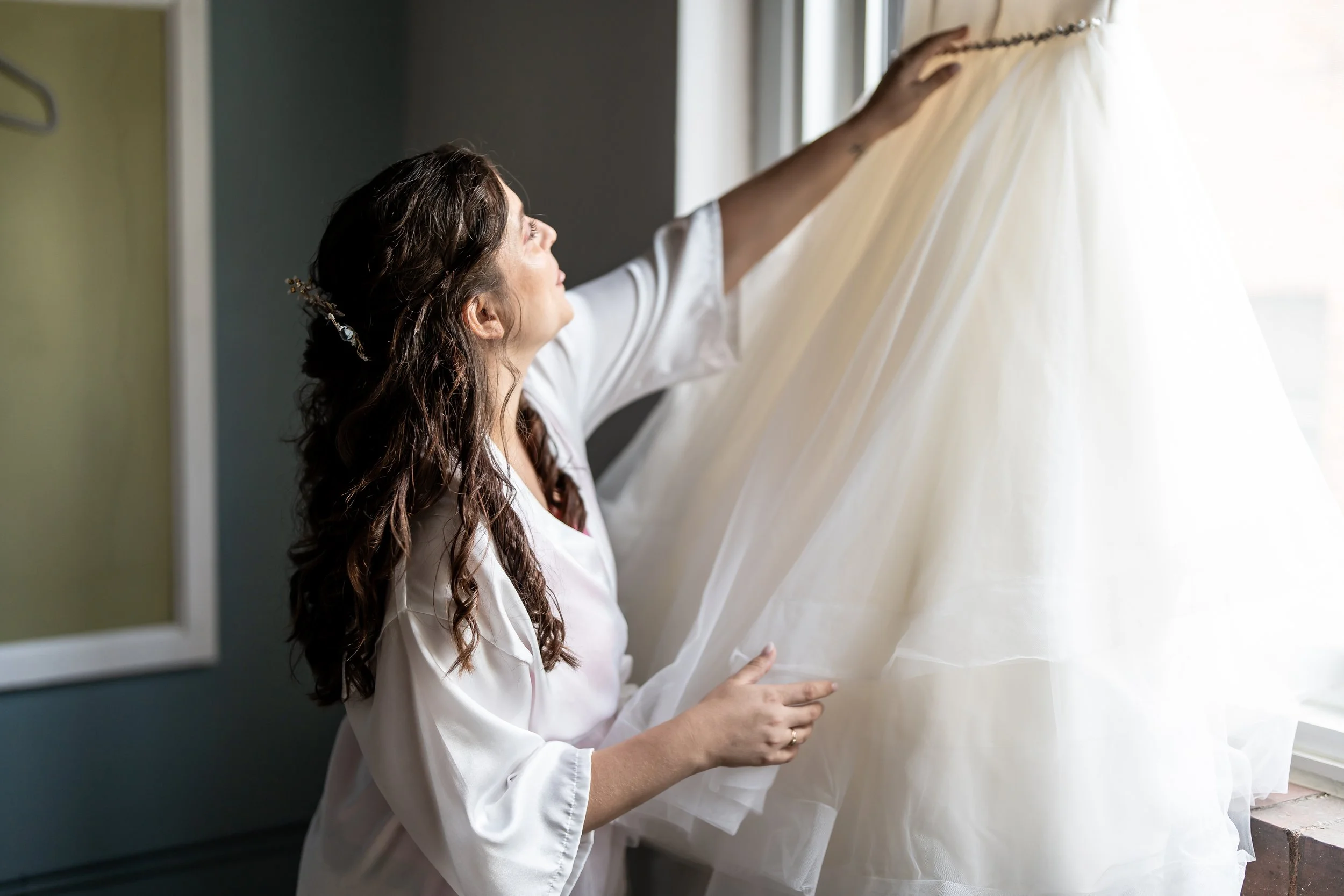 A woman with long brown hair, wearing a white satin robe, is looking at and touching a white wedding dress hanging near a window.