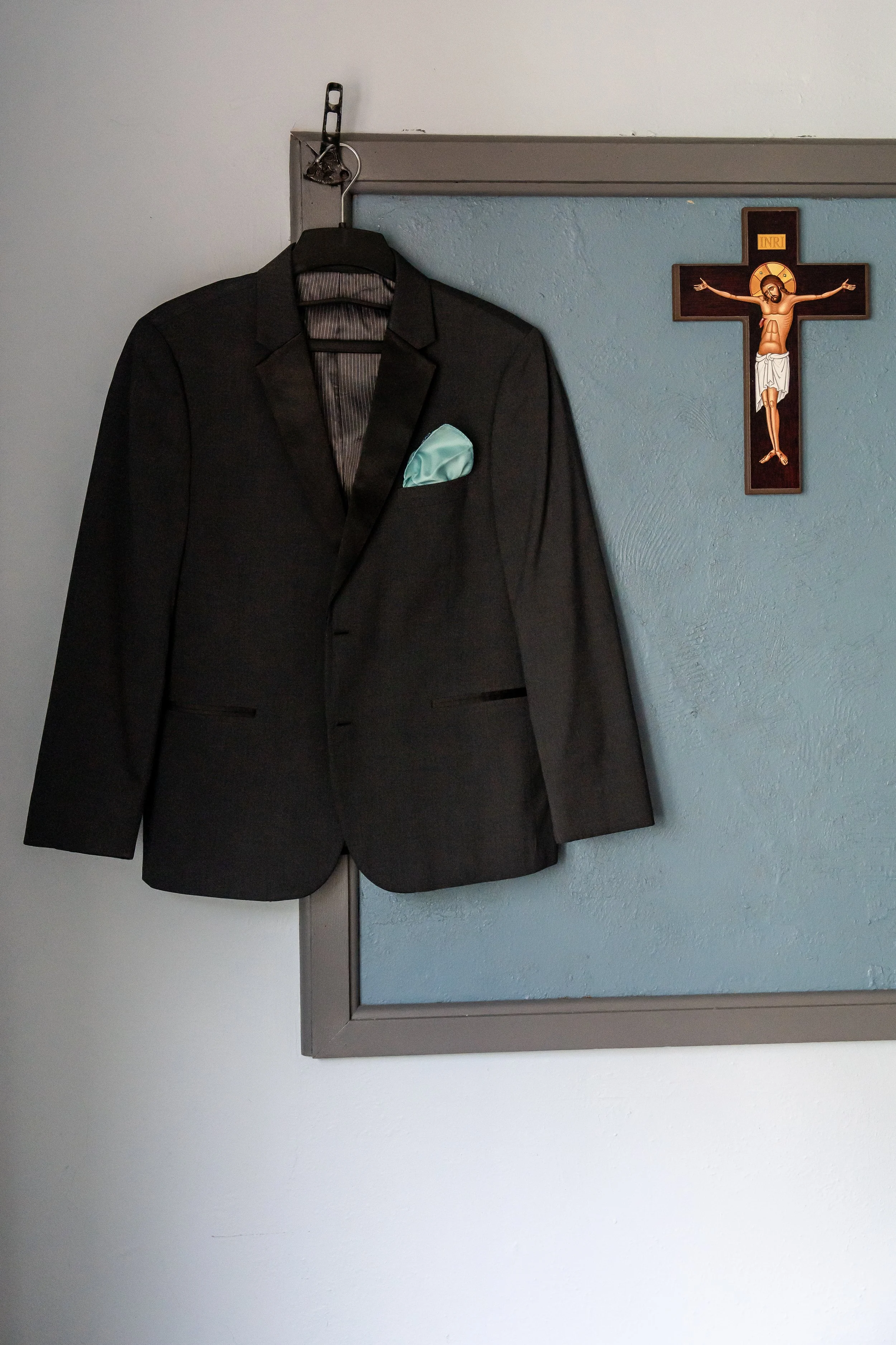 A black suit jacket with a light blue pocket square hanging on a hanger on a wall, with a small crucifix with a figure of Jesus Christ on the same wall.