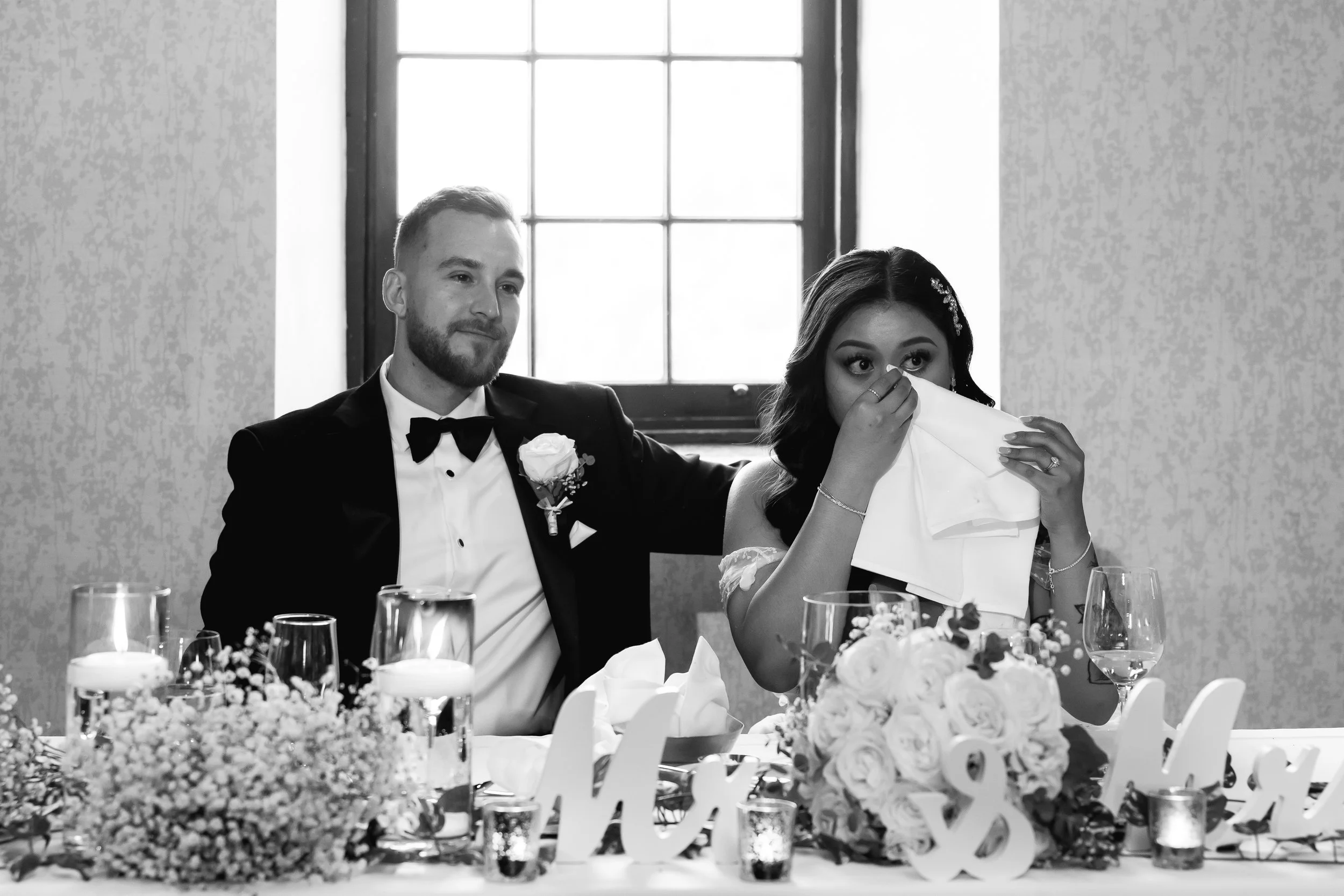 A bride and groom at their wedding reception, the groom wearing a tuxedo with a bow tie and a white boutonniere, seated next to the bride who is wiping her eyes with a napkin. The wedding table is decorated with flowers, candles, and signs, and a lar