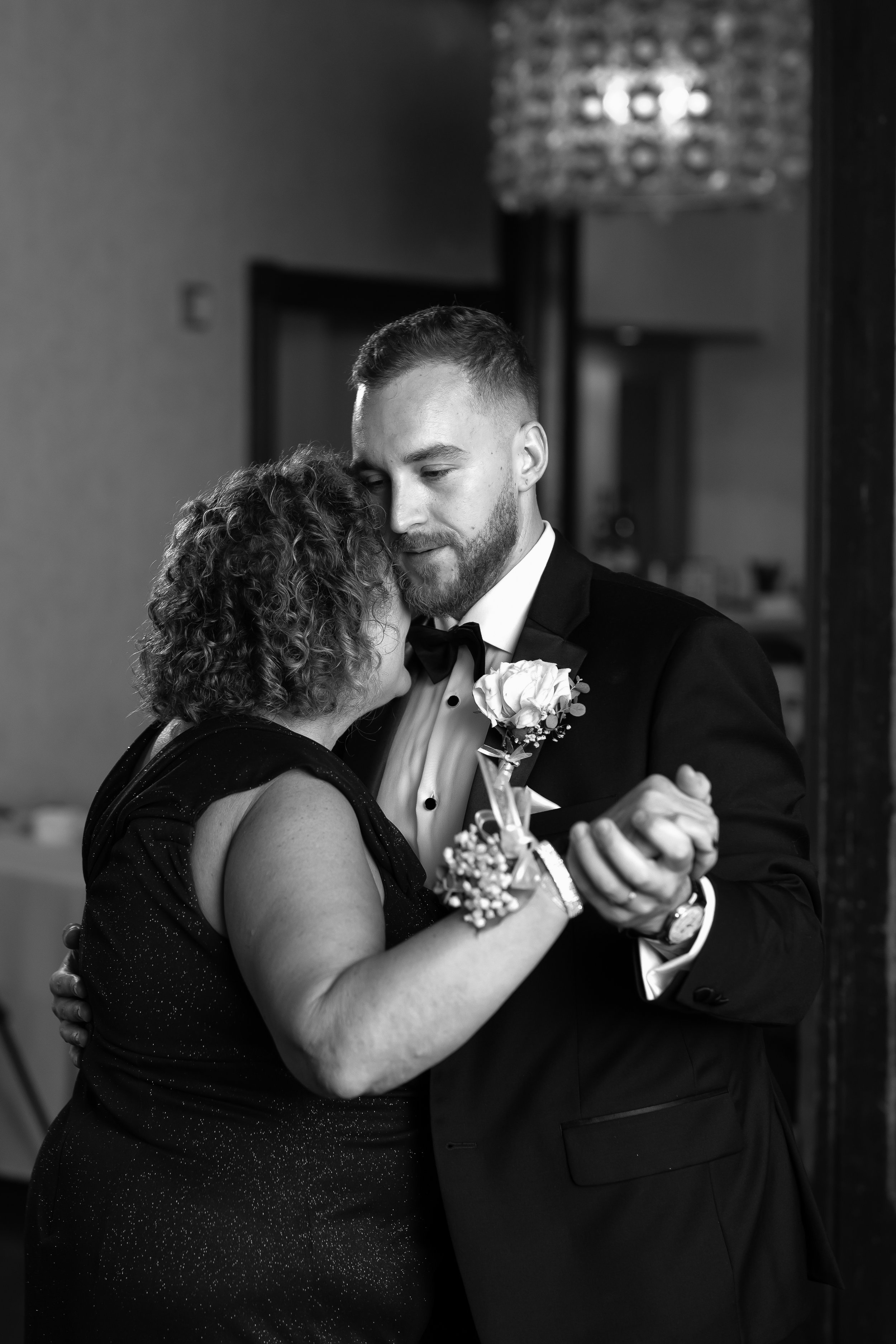 A black and white photo of a groom dancing closely with a woman, possibly his mother, at a wedding reception. The groom is wearing a tuxedo and has a boutonniere on his lapel. The woman has curly hair and is dressed in a sleeveless, sparkly dress.