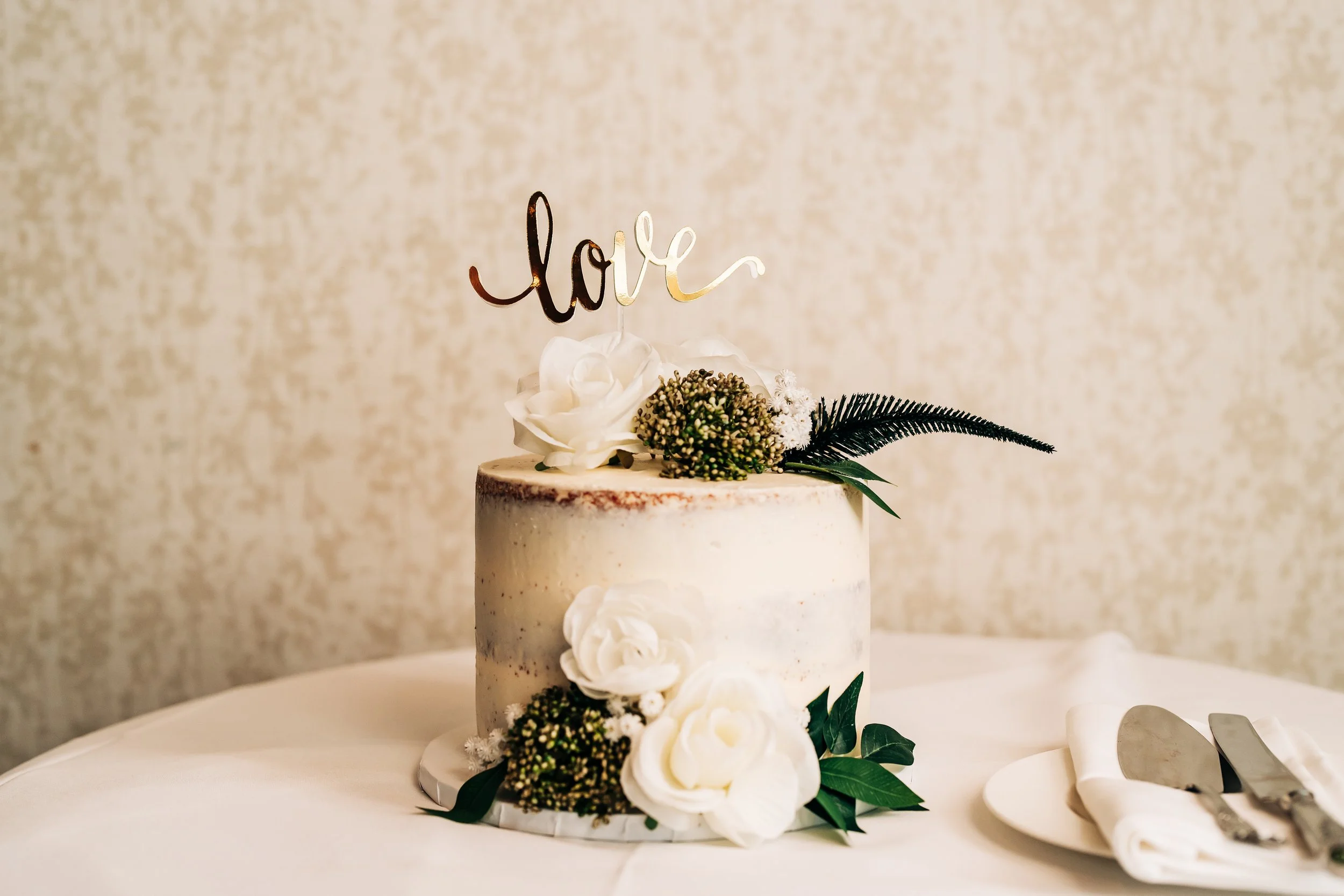 A white wedding cake decorated with white flowers and greenery, with a gold 'love' cake topper, on a white tablecloth, with a plate and cake utensils nearby.